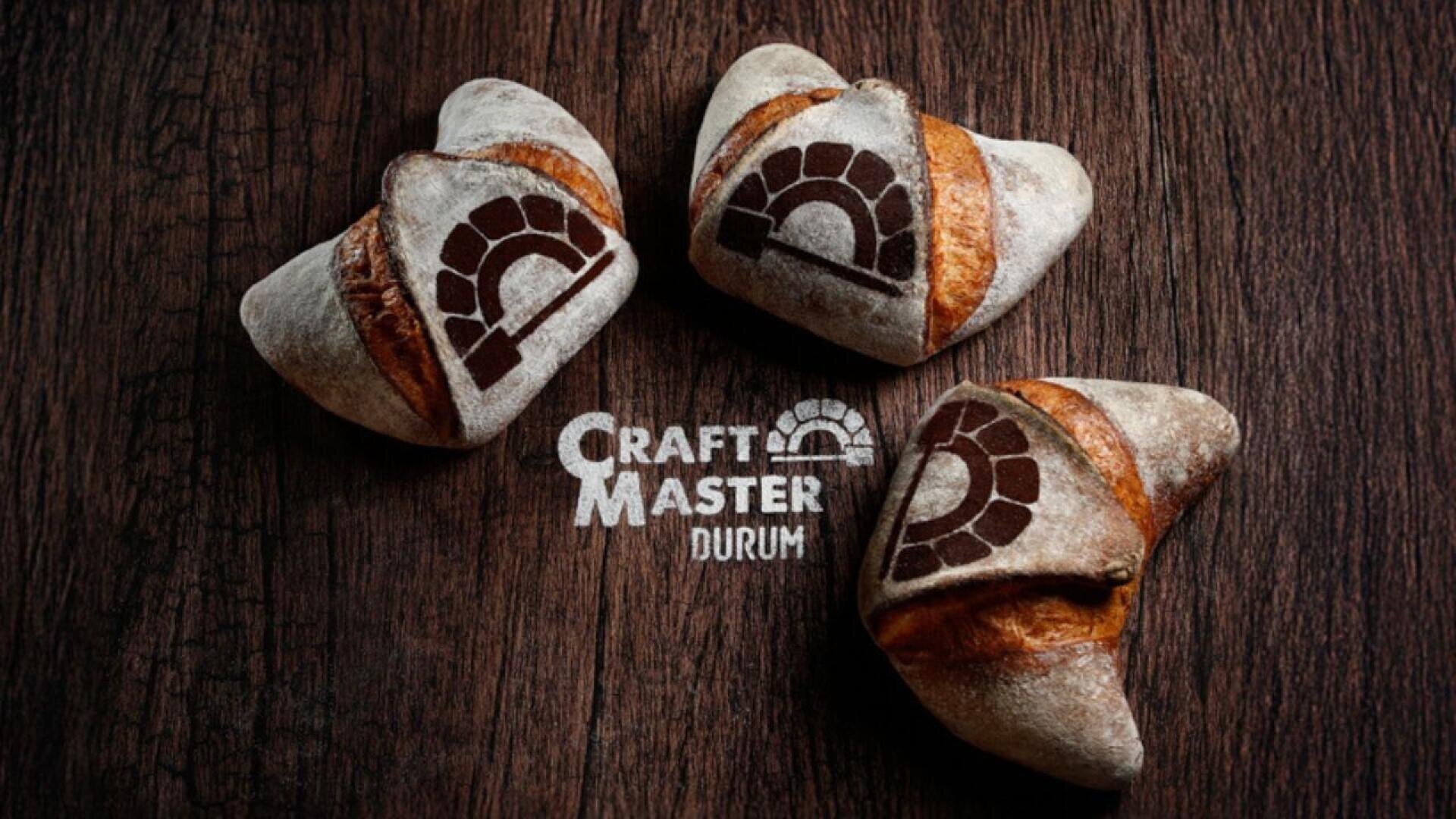 Three rustic crescent bread rolls with a dark oven logo stamped on them are arranged on a wooden surface above the text Craft Master Durum.