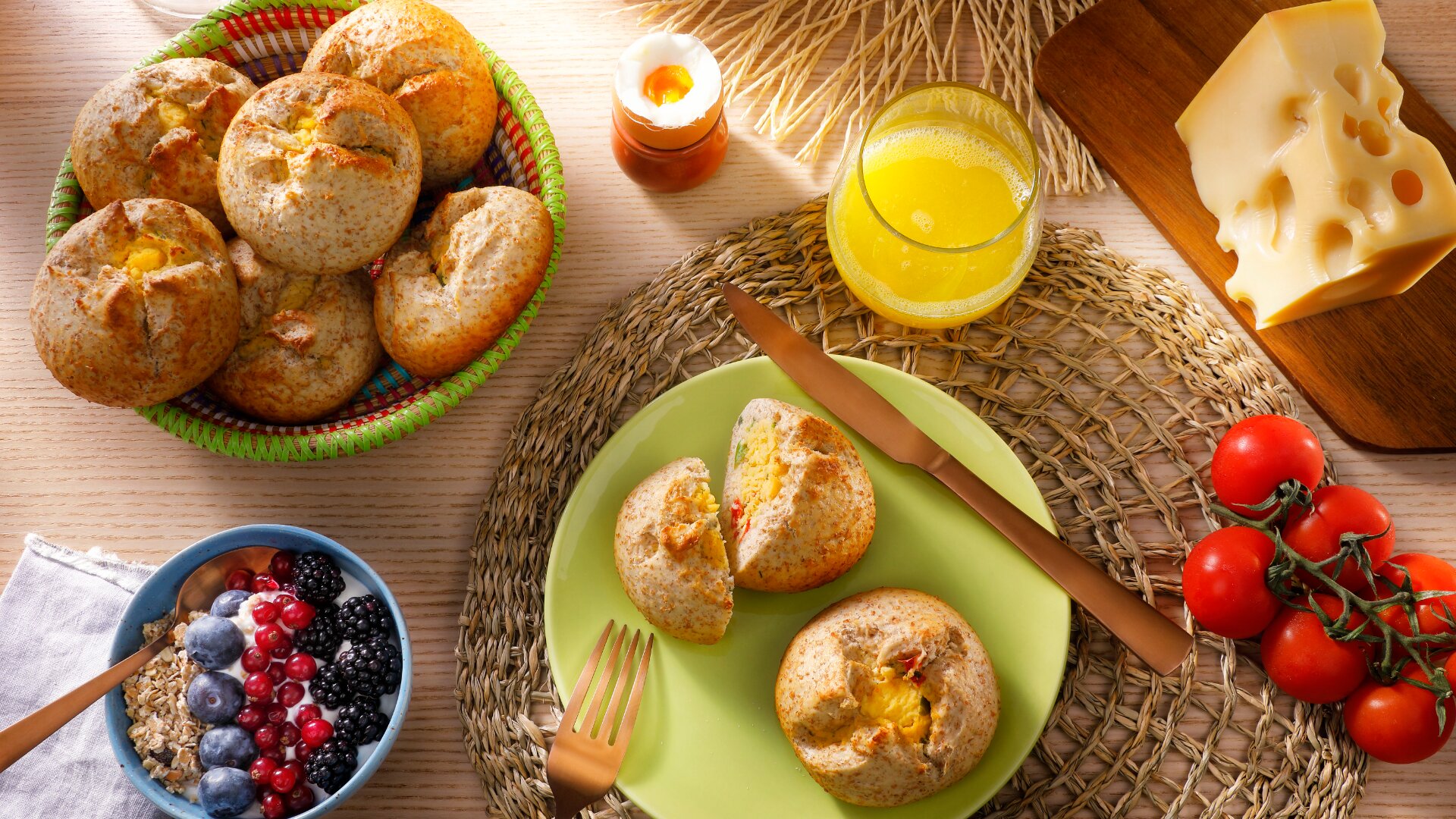 A breakfast spread with whole grain rolls, a bowl of granola topped with berries, a glass of orange juice, sliced cheese, tomatoes on the vine, and a lit candle on a woven placemat.