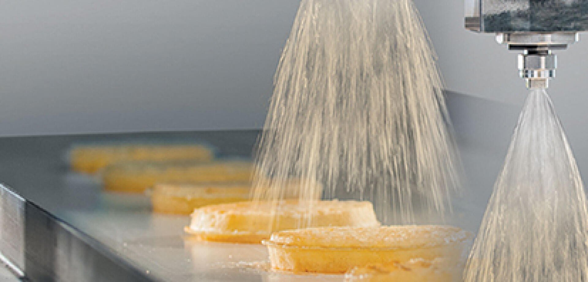 In einer industriellen Lebensmittelverarbeitungsanlage wird Zucker gleichmäßig auf rundes Gebäck gestreut, das auf einem Förderband läuft.