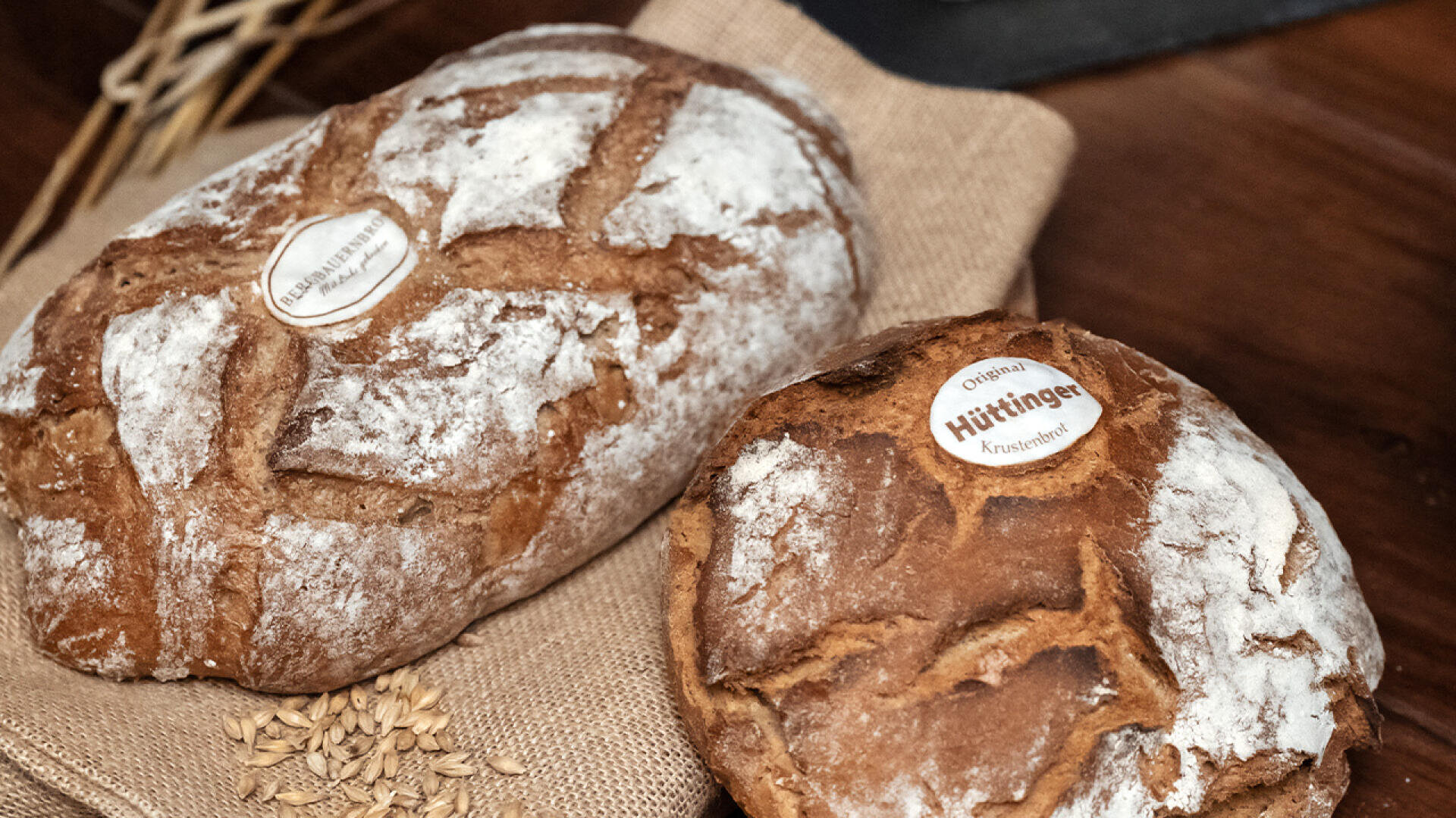 Zwei rustikale Brotlaibe mit mehlbestäubter Kruste liegen auf einem Jutetuch. Auf jedem Laib befindet sich ein kleines rundes Etikett, daneben liegen verstreut ein paar Weizenkörner.