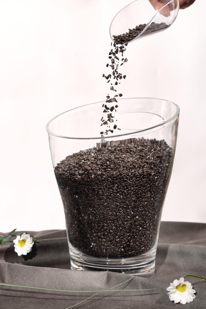 A hand pours black chia seeds from a small container into a large glass that is already nearly full of chia seeds. The glass sits on a gray surface with white flowers nearby.