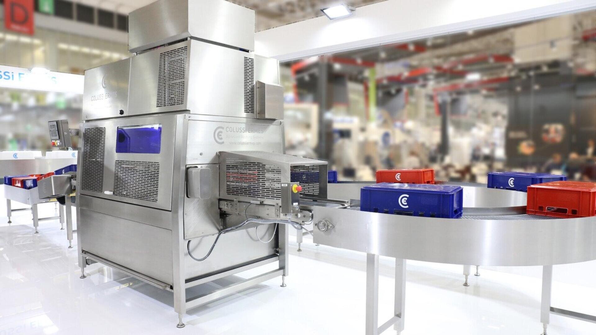 Large industrial food processing machine with conveyor belts transporting red and blue plastic crates in a bright, modern factory setting. Background shows blurred equipment and people.