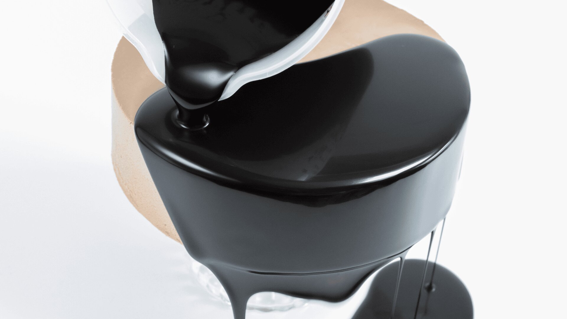 A close-up of silky black liquid being poured from a white container over a round, beige dessert, creating a glossy, smooth coating that drips down the sides onto a white surface.
