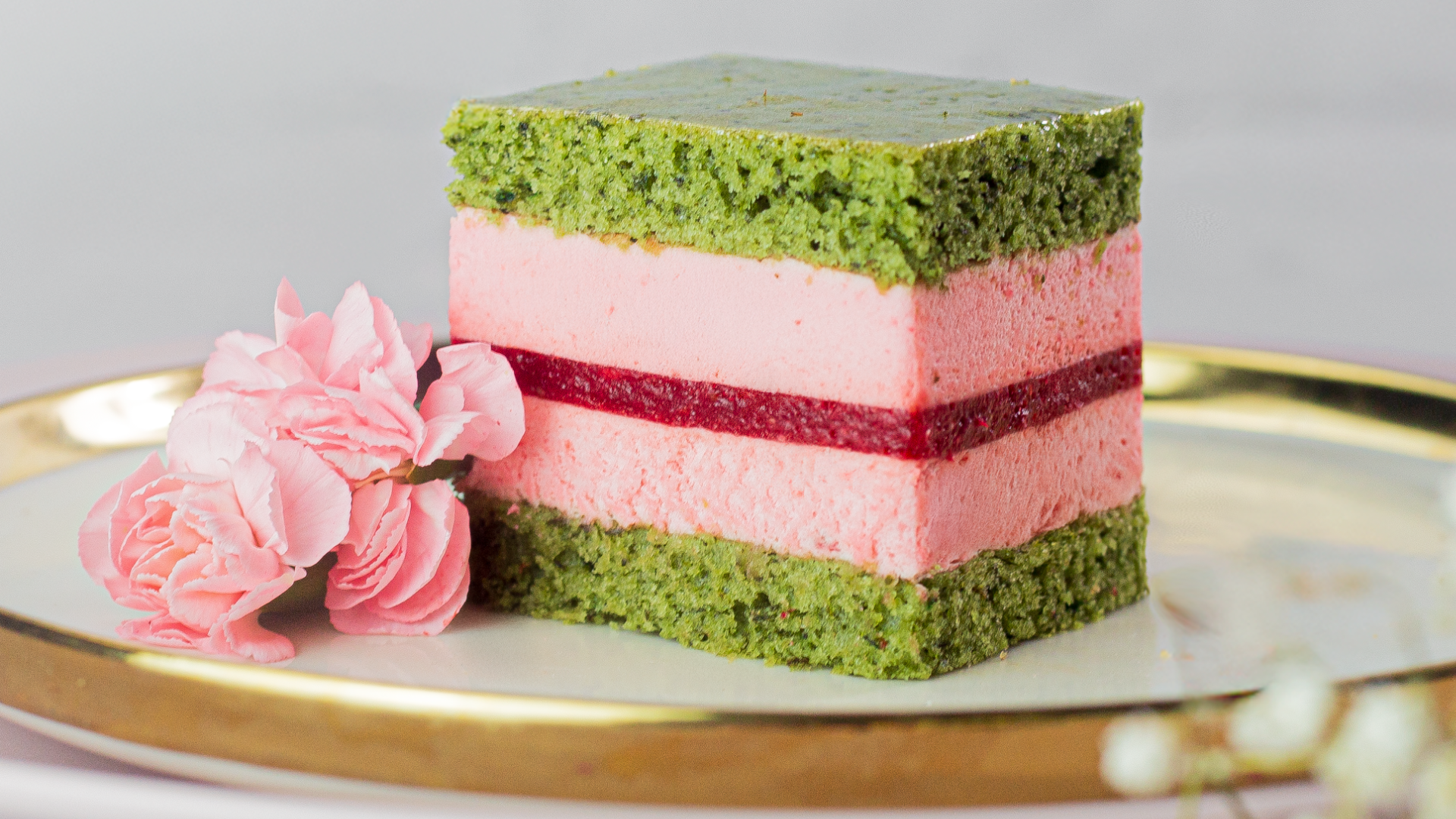 A slice of layered cake with green sponge, pink mousse, and a red jelly layer, garnished with two pink flowers, served on a gold-rimmed white plate.