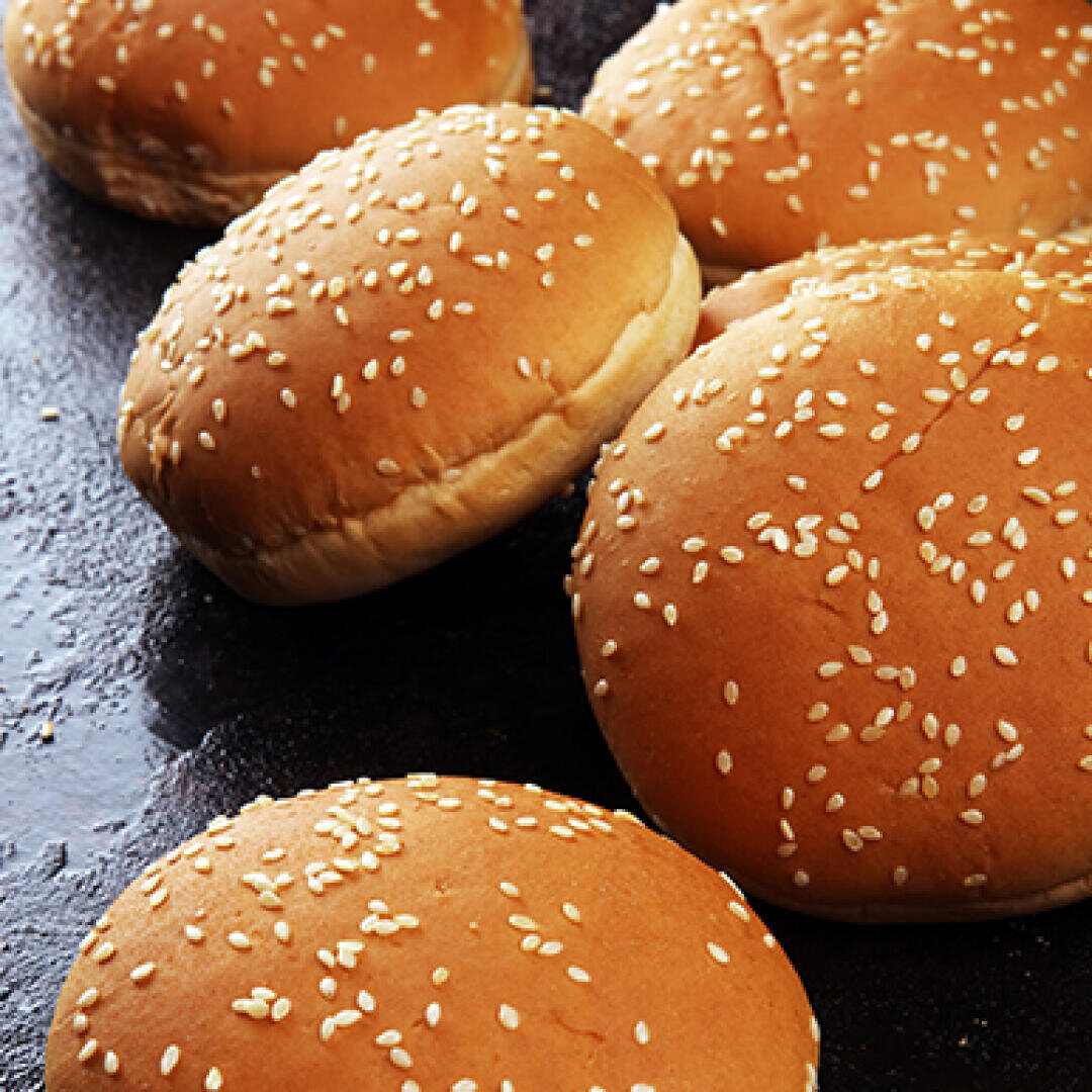 Several sesame seed hamburger buns are arranged on a dark surface, with some buns overlapping each other. The buns are golden brown and topped with scattered sesame seeds.