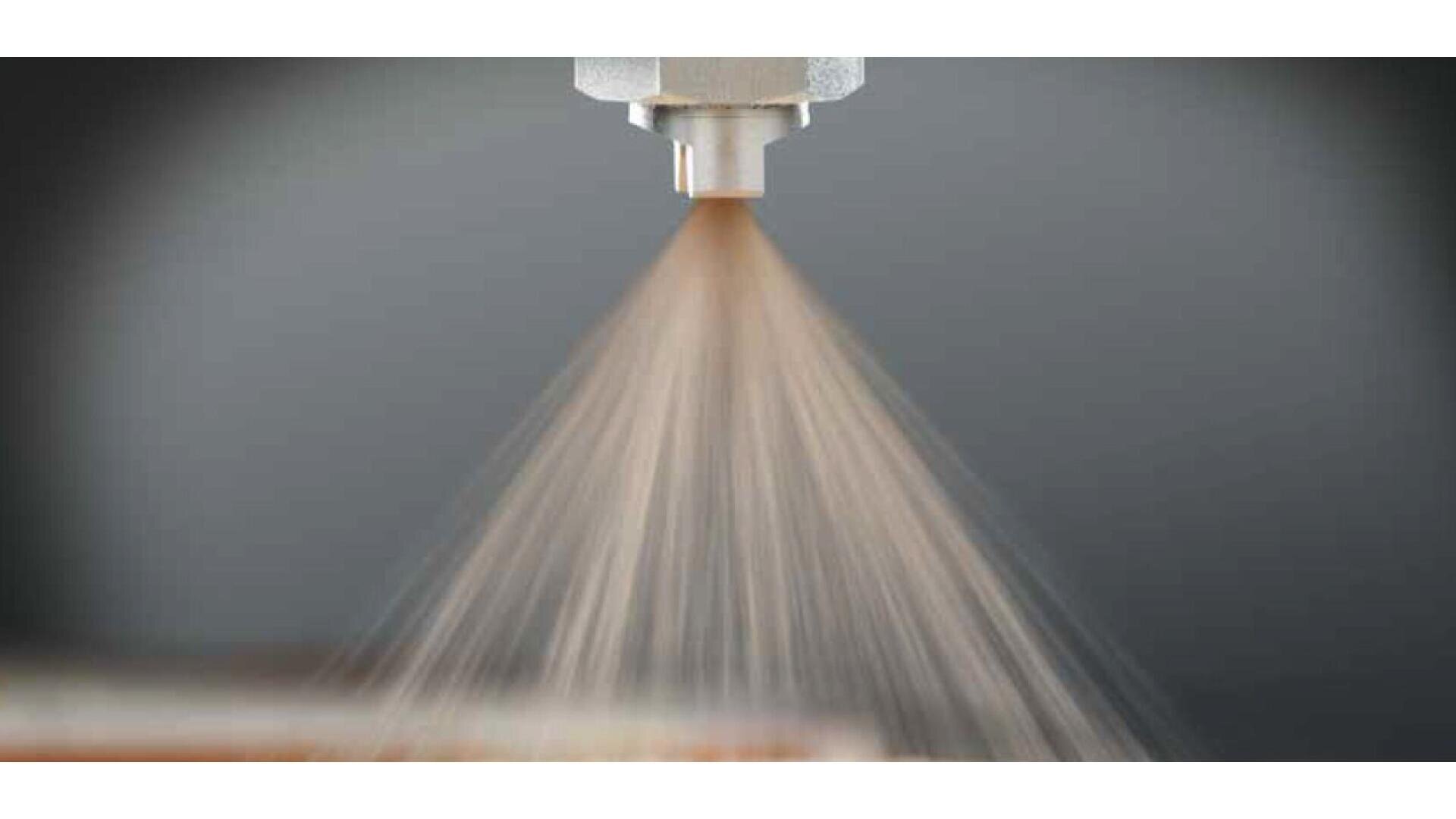 A close-up view of a spray nozzle releasing a fine mist or spray of liquid, with the mist spreading outward in a fan shape against a blurred background.