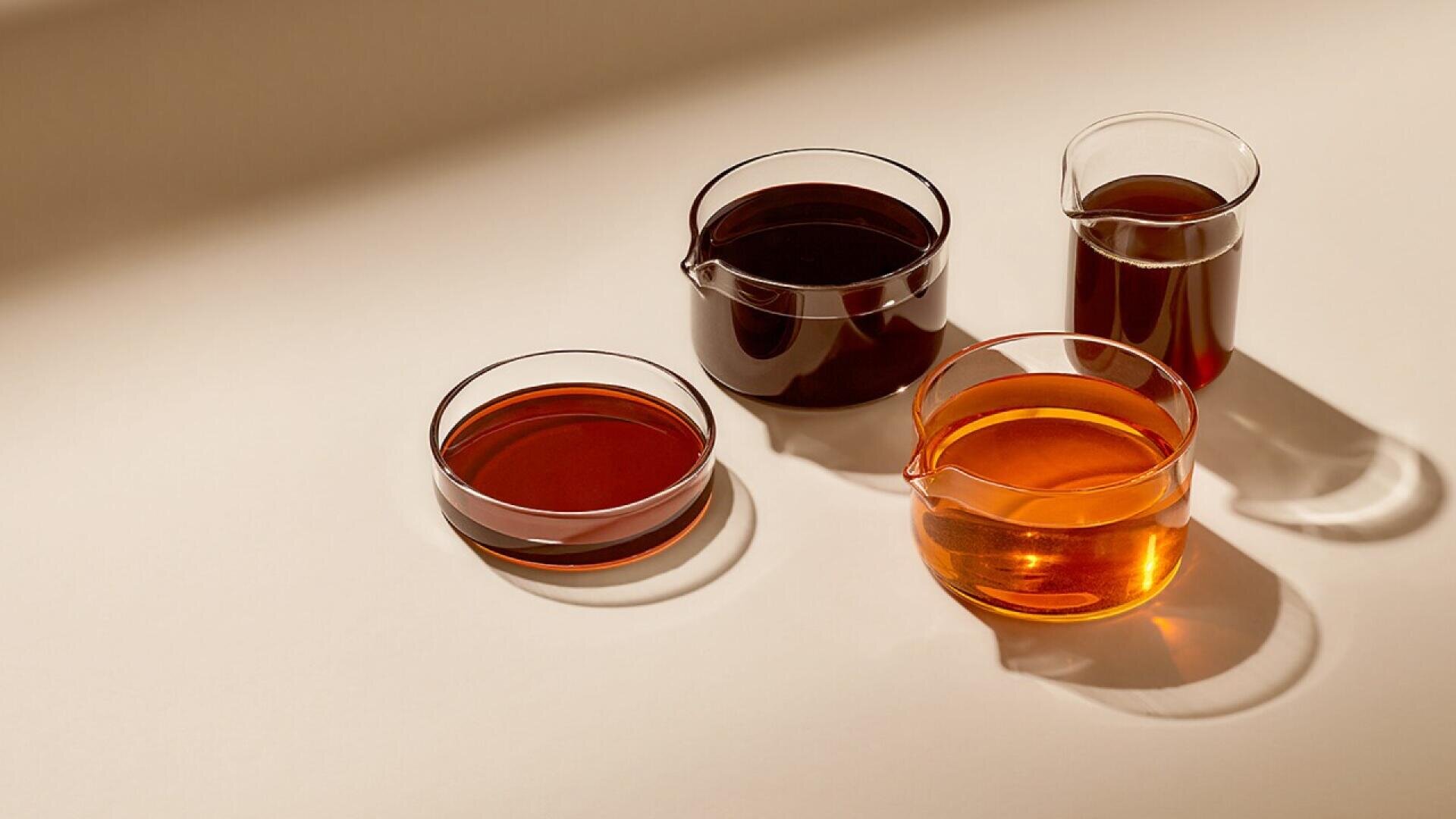 Four glass containers filled with different shades of amber and brown liquids are arranged on a neutral surface, casting soft shadows in the light.