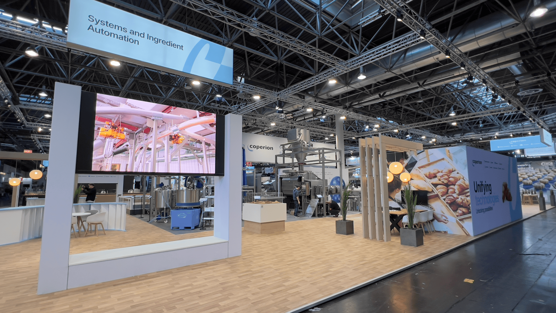 A modern trade show booth with a large screen displaying industrial equipment, seating areas, and a sign reading Systems and Ingredient Automation. Decorative lights and a wall with bread images are also visible.