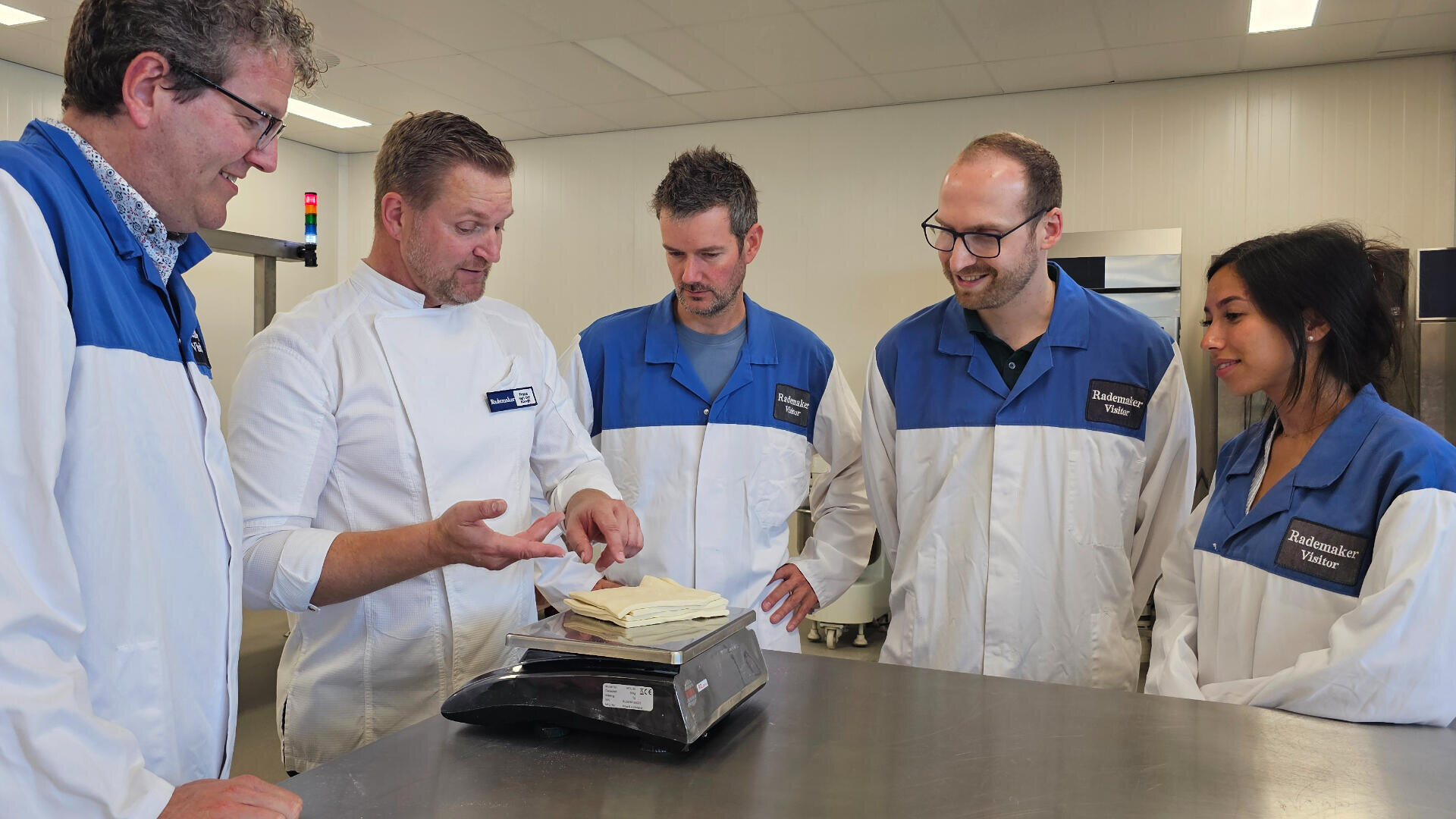 Five people in lab coats stand around a metal table, watching a man in a chefs coat demonstrate something using a block of butter on a digital scale in a bright, modern kitchen or lab setting.