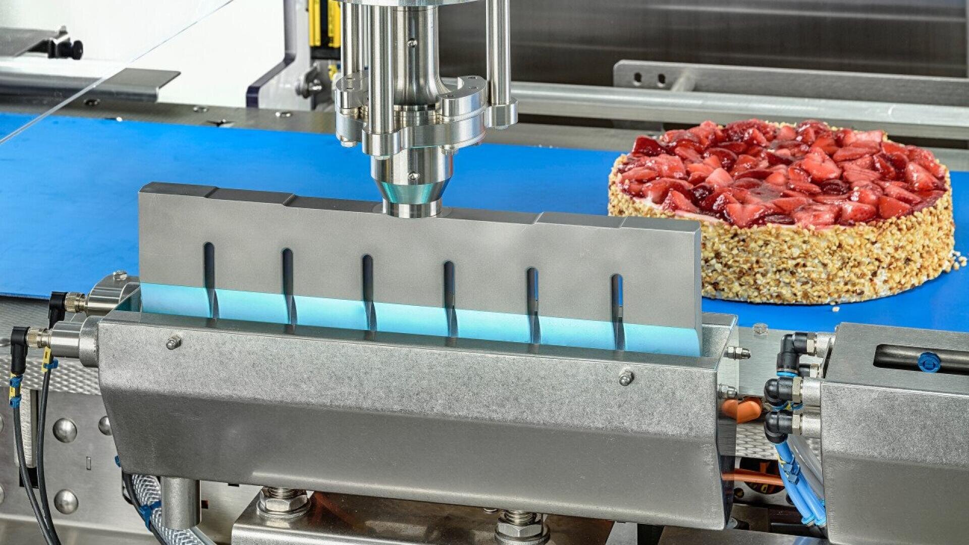 A machine with metal blades is slicing a round strawberry-topped cake with a nut-covered edge on a blue conveyor belt in a food processing facility.