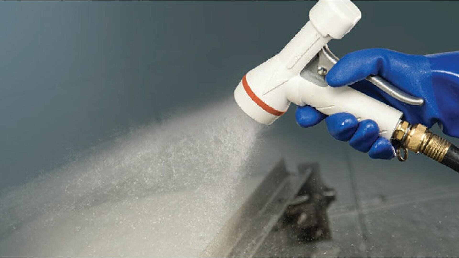 A person wearing a blue glove uses a white spray nozzle to release a fine mist of liquid over a metal surface.