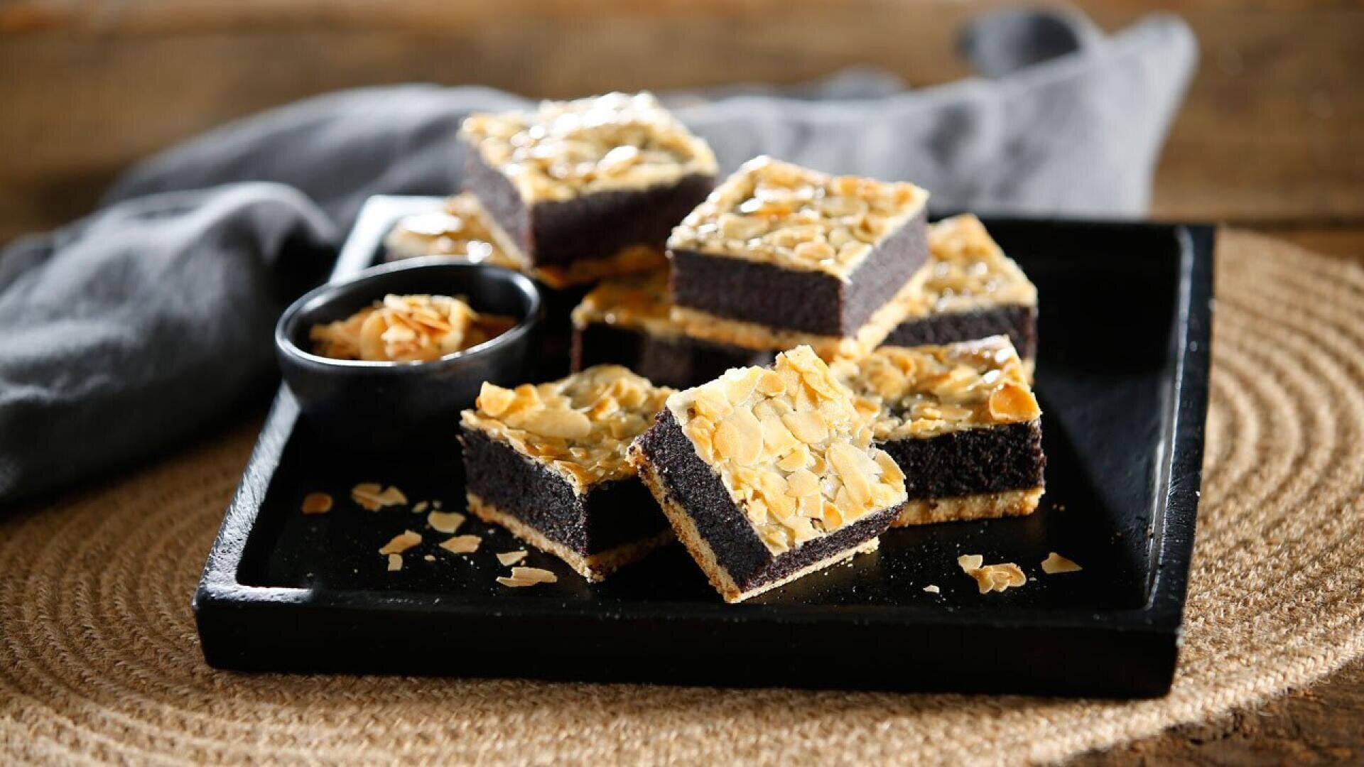 Squares of layered poppy seed cake topped with sliced almonds are arranged on a black tray, with a small black bowl of extra almonds and a gray cloth napkin in the background.