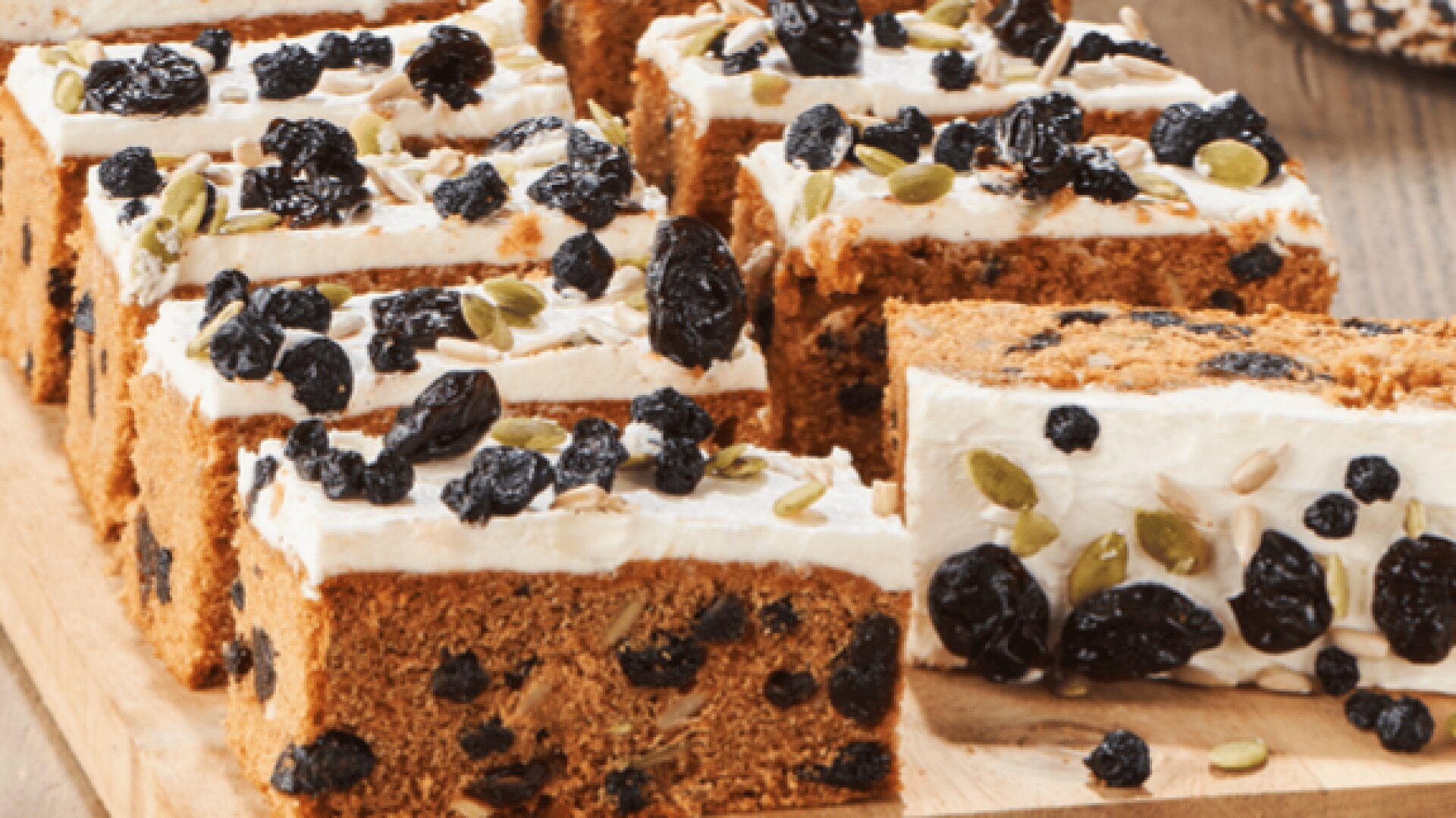 Sliced pieces of cake with white frosting, topped with dried berries and pumpkin seeds, arranged on a wooden board.