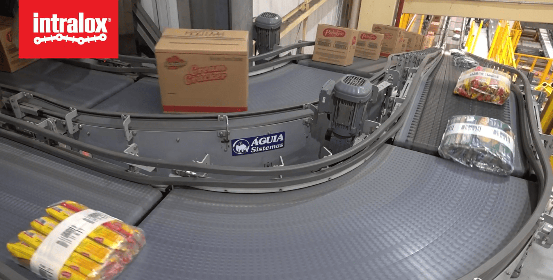 Conveyor belts transport packaged food items and cardboard boxes through a factory. The Intralox logo appears in the top left corner, and an Aguta Sistemas sign is visible on the conveyor system.