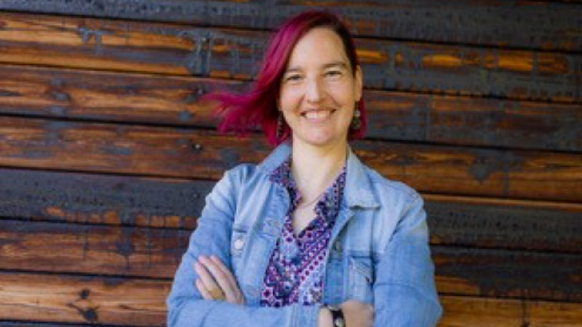Eine Person mit kurzen rosa Haaren lächelt mit verschränkten Armen, trägt eine Jeansjacke über einem gemusterten Hemd und steht vor einer Holzwand.