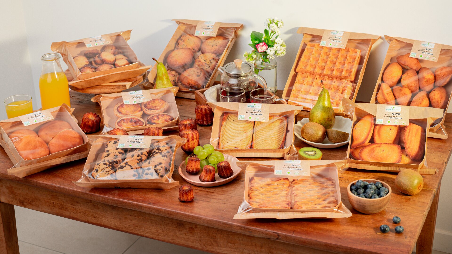 A wooden table filled with various packaged baked goods, fresh fruits, two glasses and a bottle of orange juice, two jars of jam, and a small flower arrangement in the background.