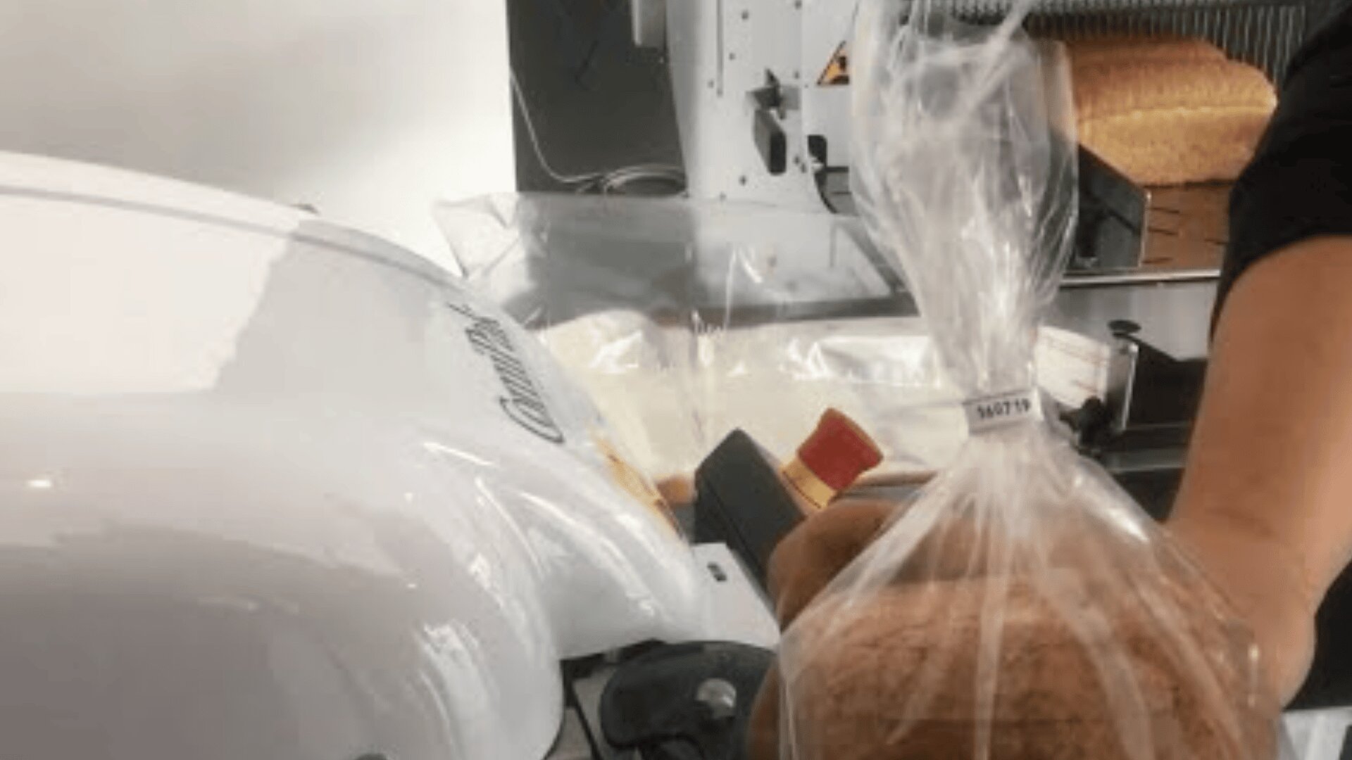 A person is holding a clear plastic bag containing a loaf of sliced bread near a bread slicing machine in a bakery or kitchen setting.