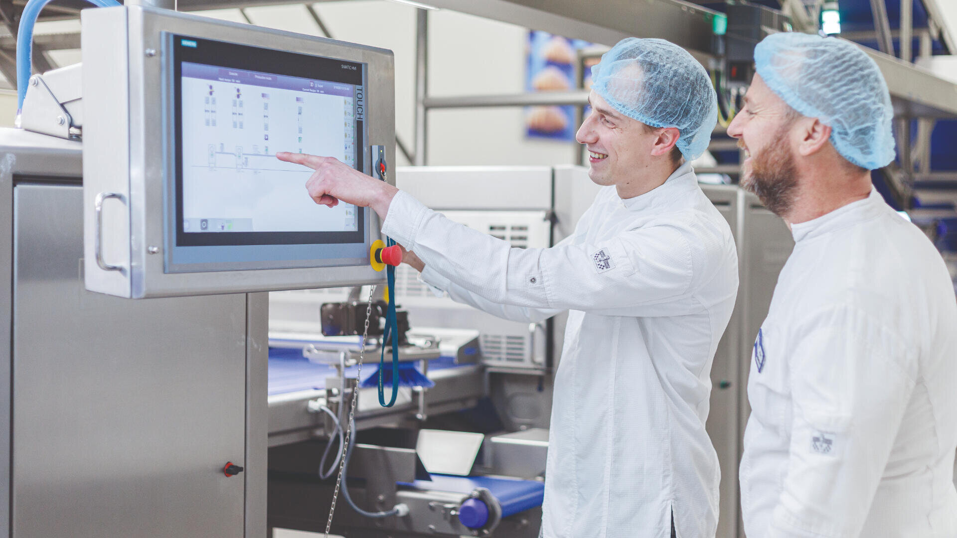Two people in white lab coats and hairnets stand in a food processing facility, looking at and pointing to a large touchscreen control panel displaying a diagram or data.