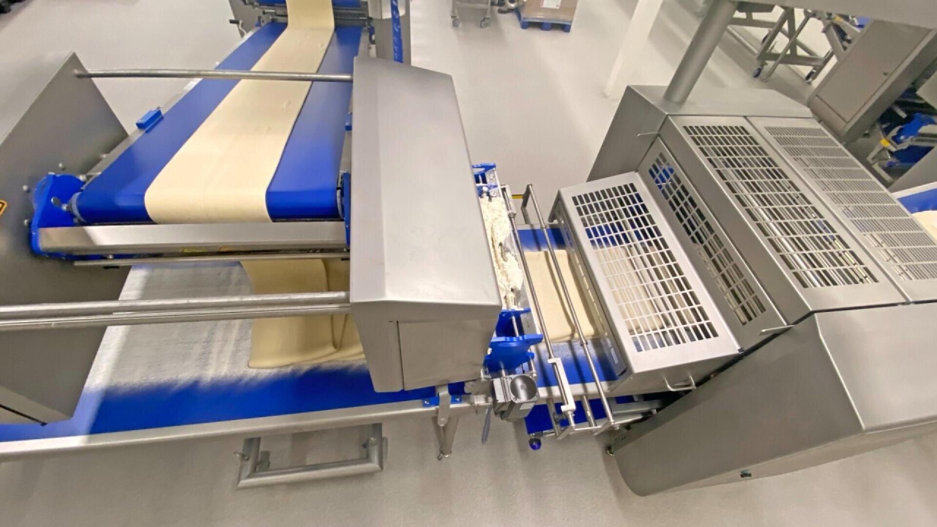 Eine handelsübliche Teigausrollmaschine in einem Lebensmittelverarbeitungsbetrieb verteilt und glättet Teigblätter auf einem blauen Förderband, das von Edelstahlkomponenten umgeben ist.