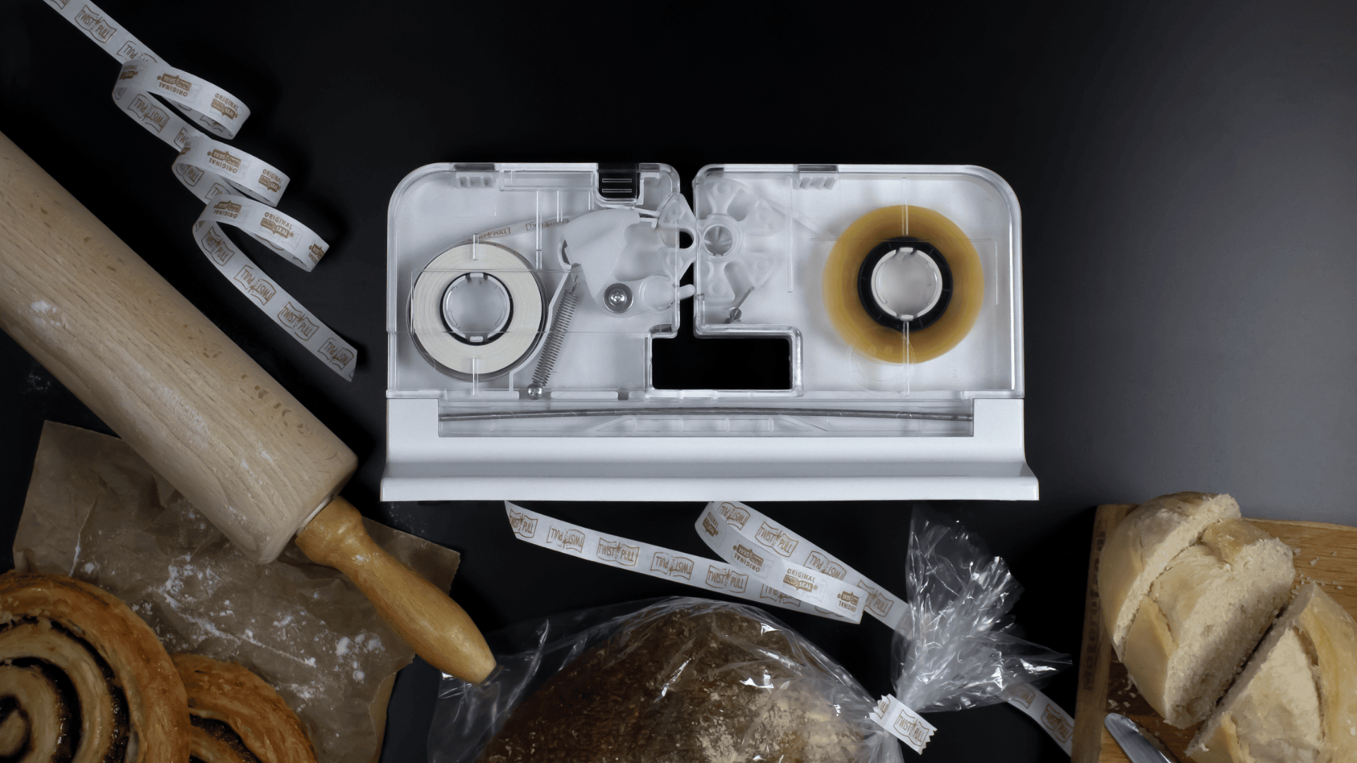 Eine Tisch-Etikettiermaschine steht auf einer schwarzen Fläche, umgeben von einem Nudelholz, Gebäck, geschnittenem Brot und Brot in einer durchsichtigen Plastiktüte. In die Maschine werden zwei Etikettenrollen eingelegt, neben denen bedruckte Etiketten liegen.