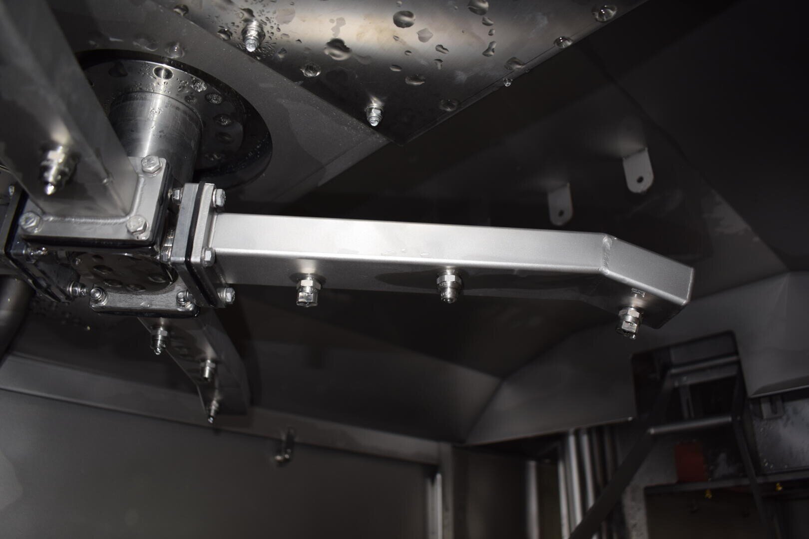 Nahaufnahme eines metallischen industriellen Maschinenteils, das einen horizontalen Metallarm mit Schrauben und andere Metallflächen mit Schrauben und Nieten zeigt, alles in einer dunklen, reflektierenden Umgebung.