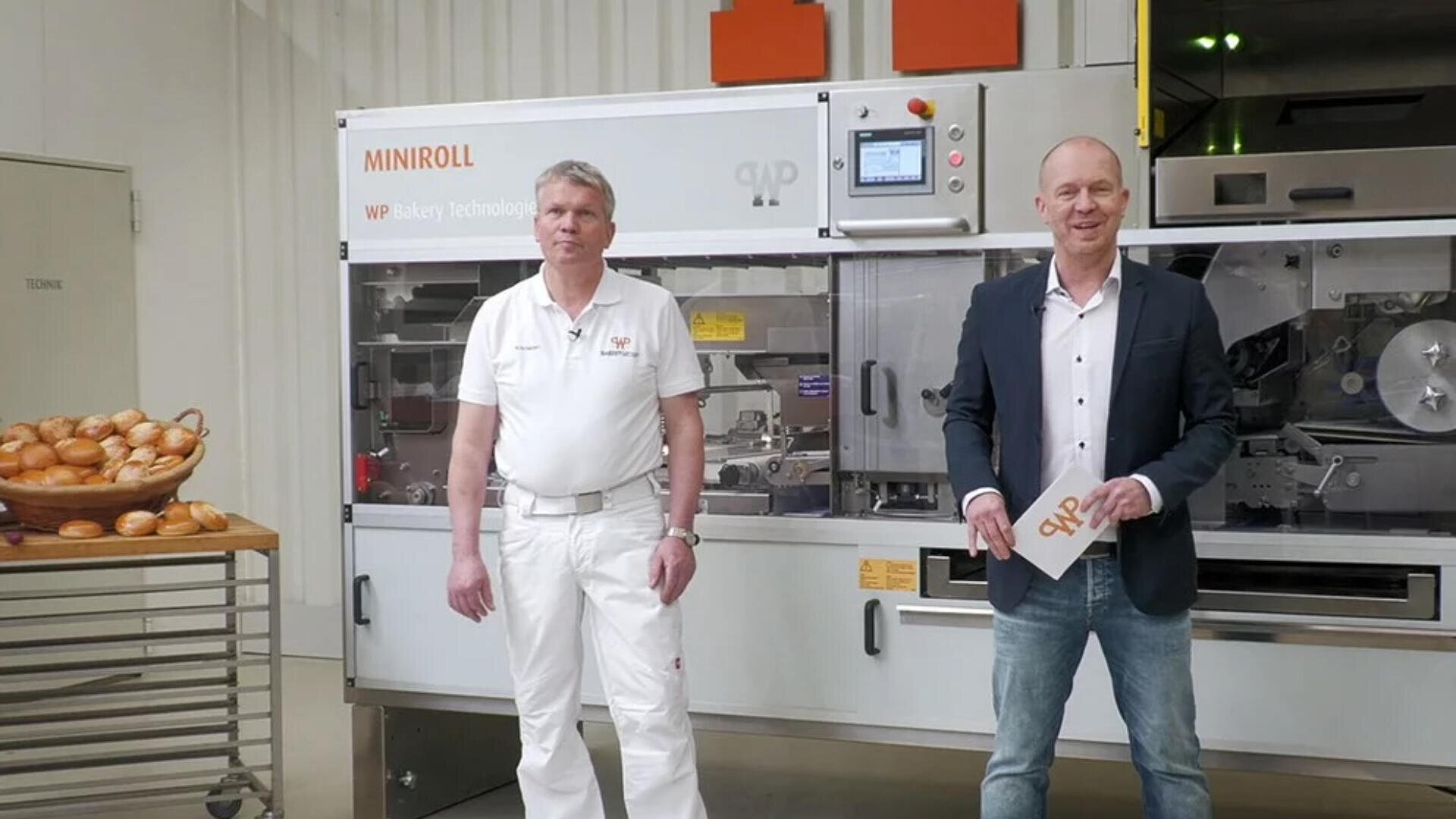Zwei Männer stehen vor einer großen Brotbackmaschine mit der Aufschrift MINIROLL. Ein Mann trägt weiße Bäckerarbeitskleidung, der andere einen Blazer und Jeans. Ein Korb mit Brötchen steht auf einem Tisch neben ihnen.