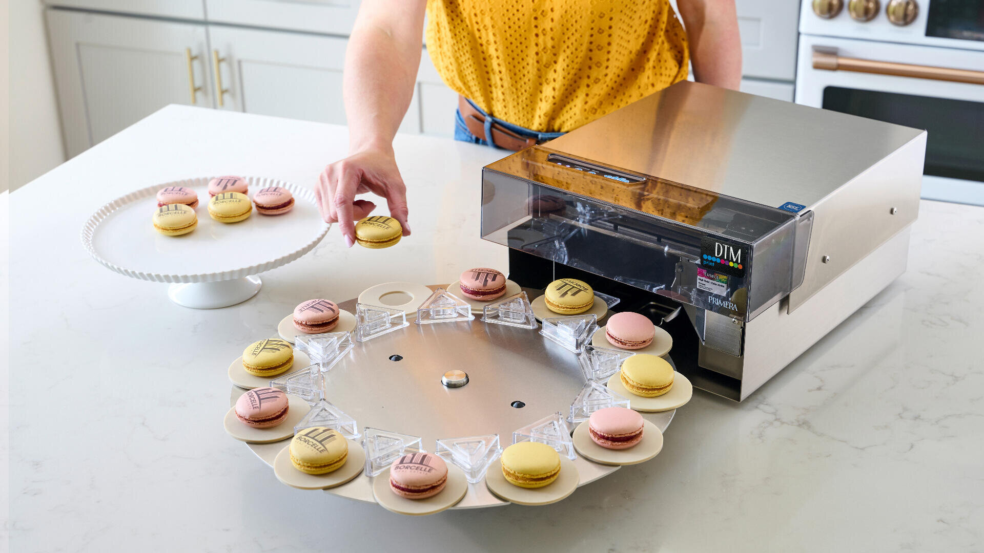 Eine Person legt ein gelbes Macaron auf einen Tortenständer, während eine Maschine in der Nähe essbare Motive auf andere Macarons druckt, die in einem Kreis auf einem rotierenden Tablett angeordnet sind. Die Szene spielt sich auf einer modernen weißen Küchentheke ab.