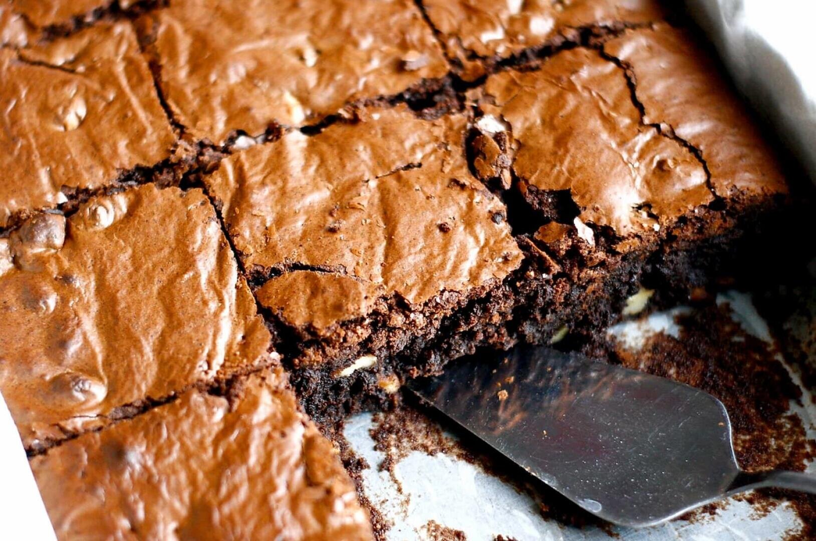 A close-up of a tray of freshly baked, fudgy chocolate brownies cut into squares, with a metal spatula lifting one piece out. The brownies have a shiny, crackly top and visible chocolate chips.