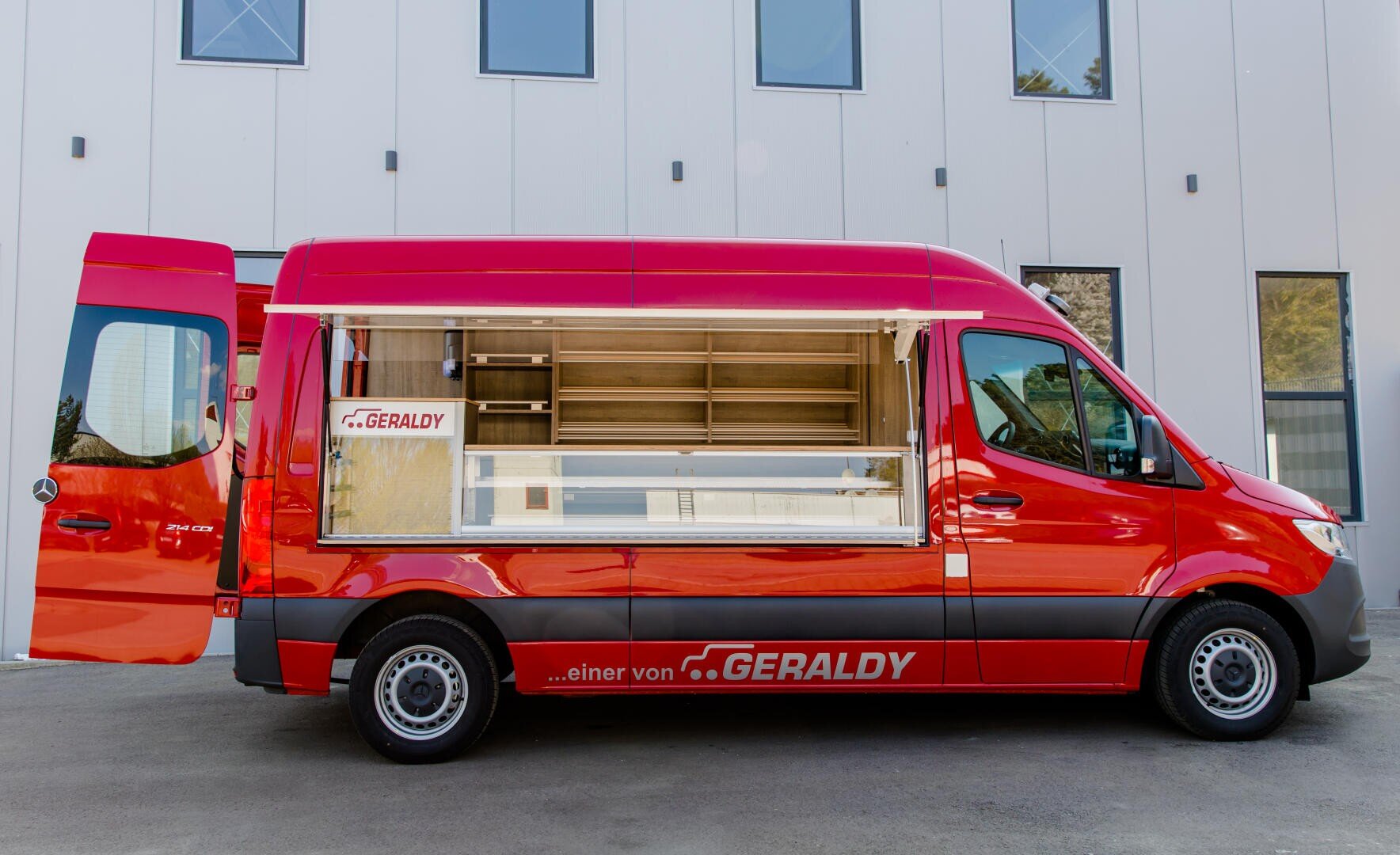 Ein leuchtend roter Lebensmitteltransporter mit großen Seiten- und Hecktüren, die sich öffnen und leere Regale im Inneren zeigen. Der Lastwagen ist vor einem modernen Gebäude mit hohen vertikalen Fenstern geparkt. Der Text "GERALDY" ist auf der Seite aufgedruckt.