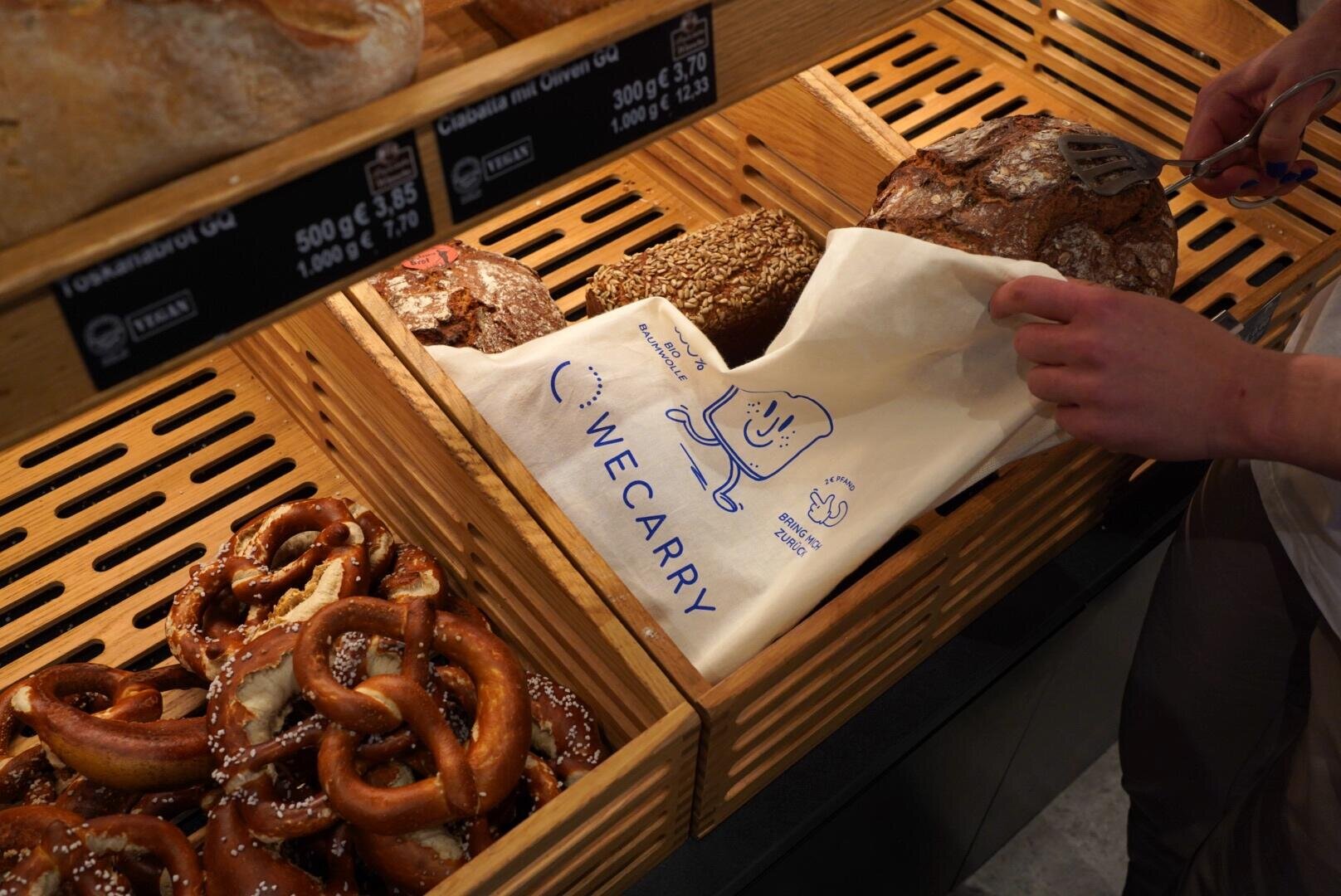 A person uses tongs to place a round loaf of bread into a cloth bag with a blue “We Carry” design at a bakery counter, surrounded by assorted bread and pretzels in wooden trays.