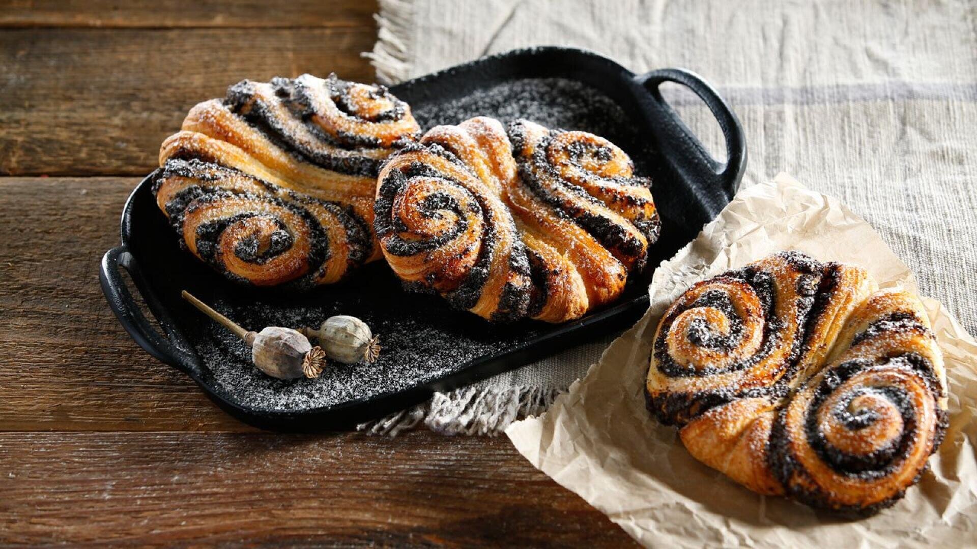 Three spiral-shaped poppy seed pastries dusted with powdered sugar are displayed on a black tray and parchment paper, set on a rustic wooden surface with a striped cloth and dried poppy pods nearby.