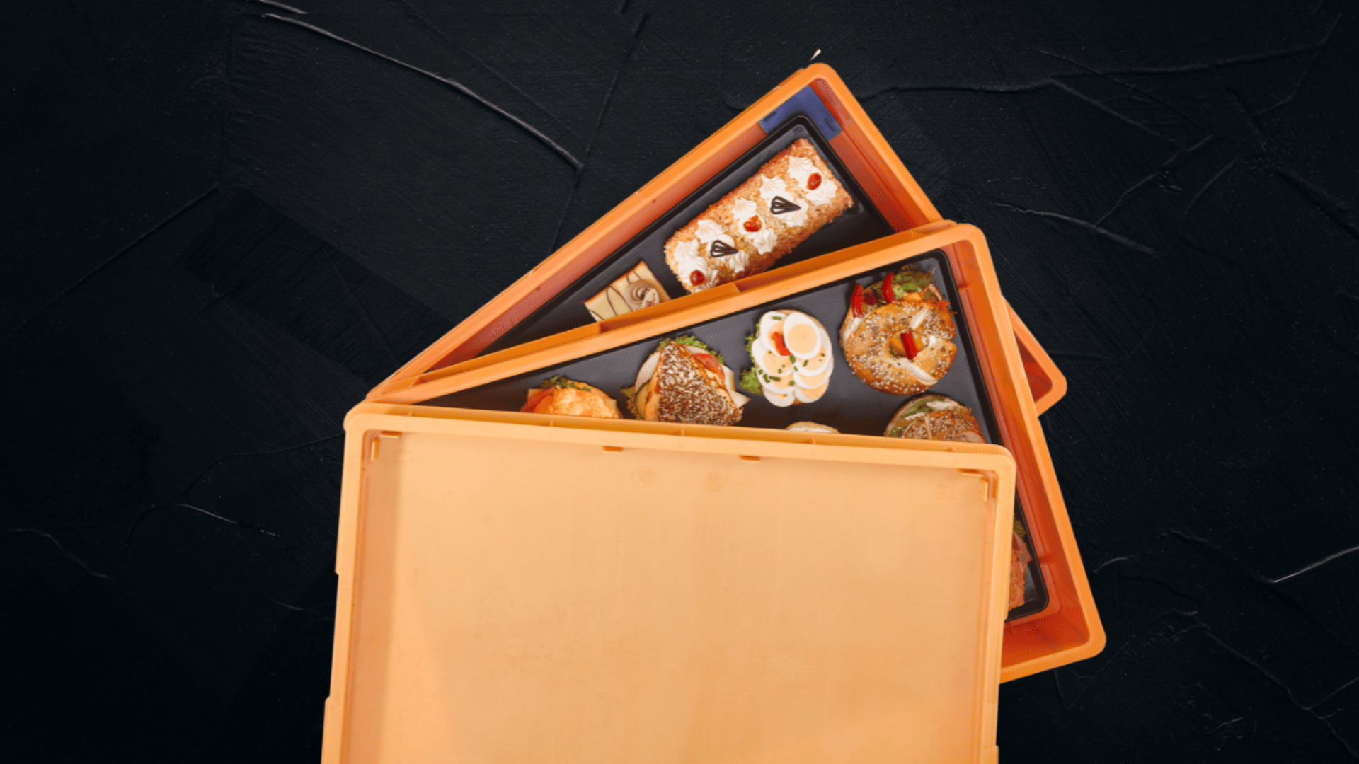 Ein orangefarbener Lebensmittelbehälter mit teilweise geöffnetem Deckel, der verschiedene Backwaren und Desserts offenbart, steht vor einem strukturierten schwarzen Hintergrund.