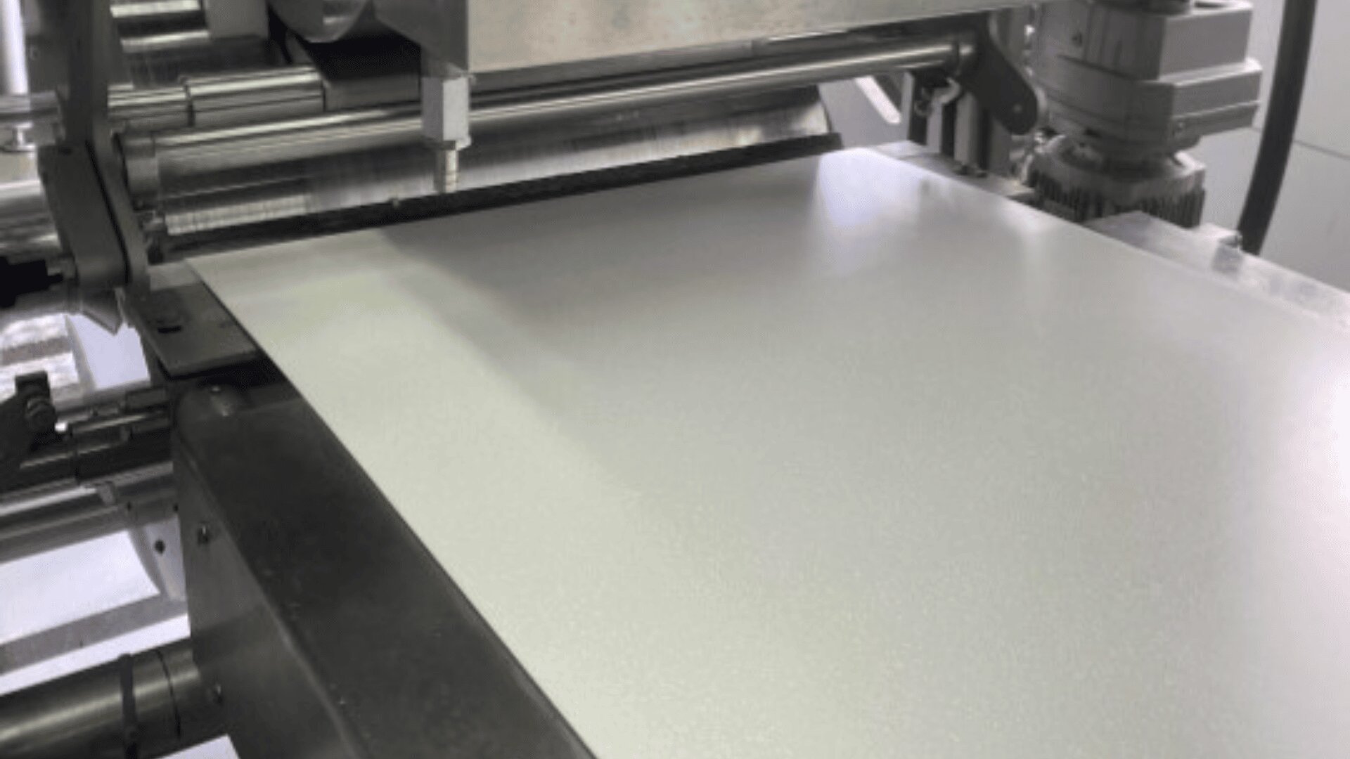 A large industrial machine processing a smooth, flat sheet of pale material, possibly metal or plastic, in a factory or manufacturing setting.