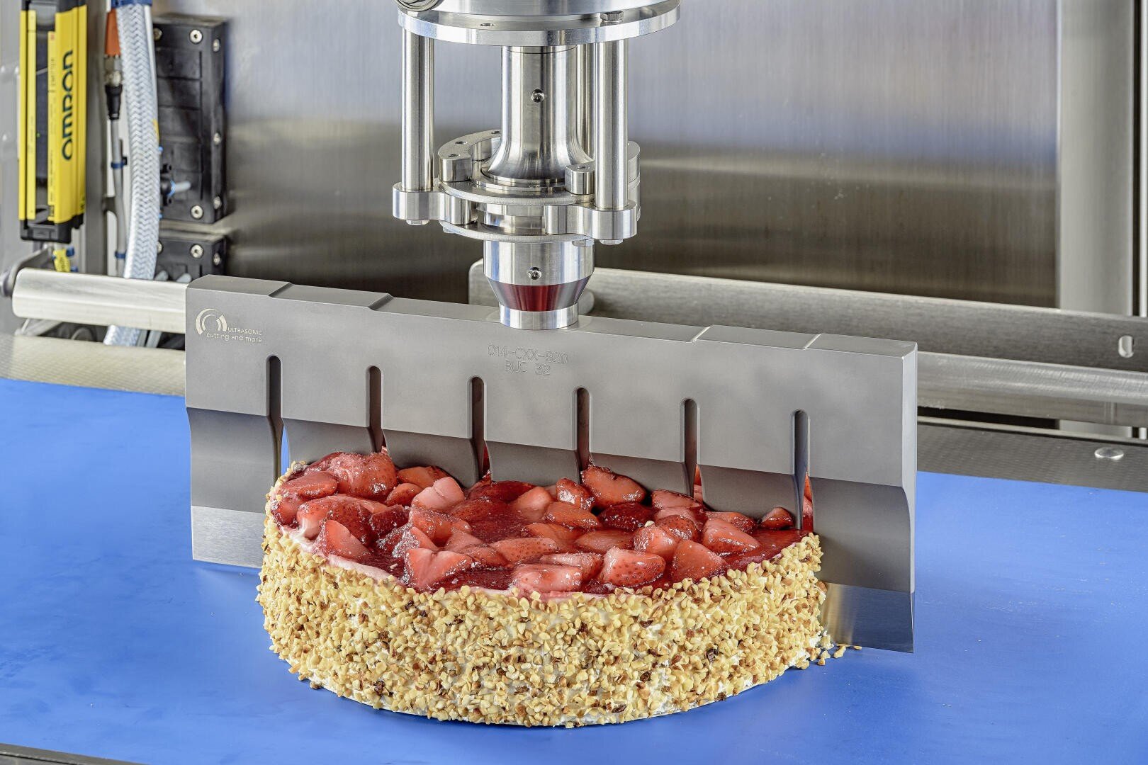 Eine Maschine mit einer Metallklinge schneidet einen runden Erdbeerkuchen mit Nussrand auf einer blauen Fläche in einer Industrieumgebung.