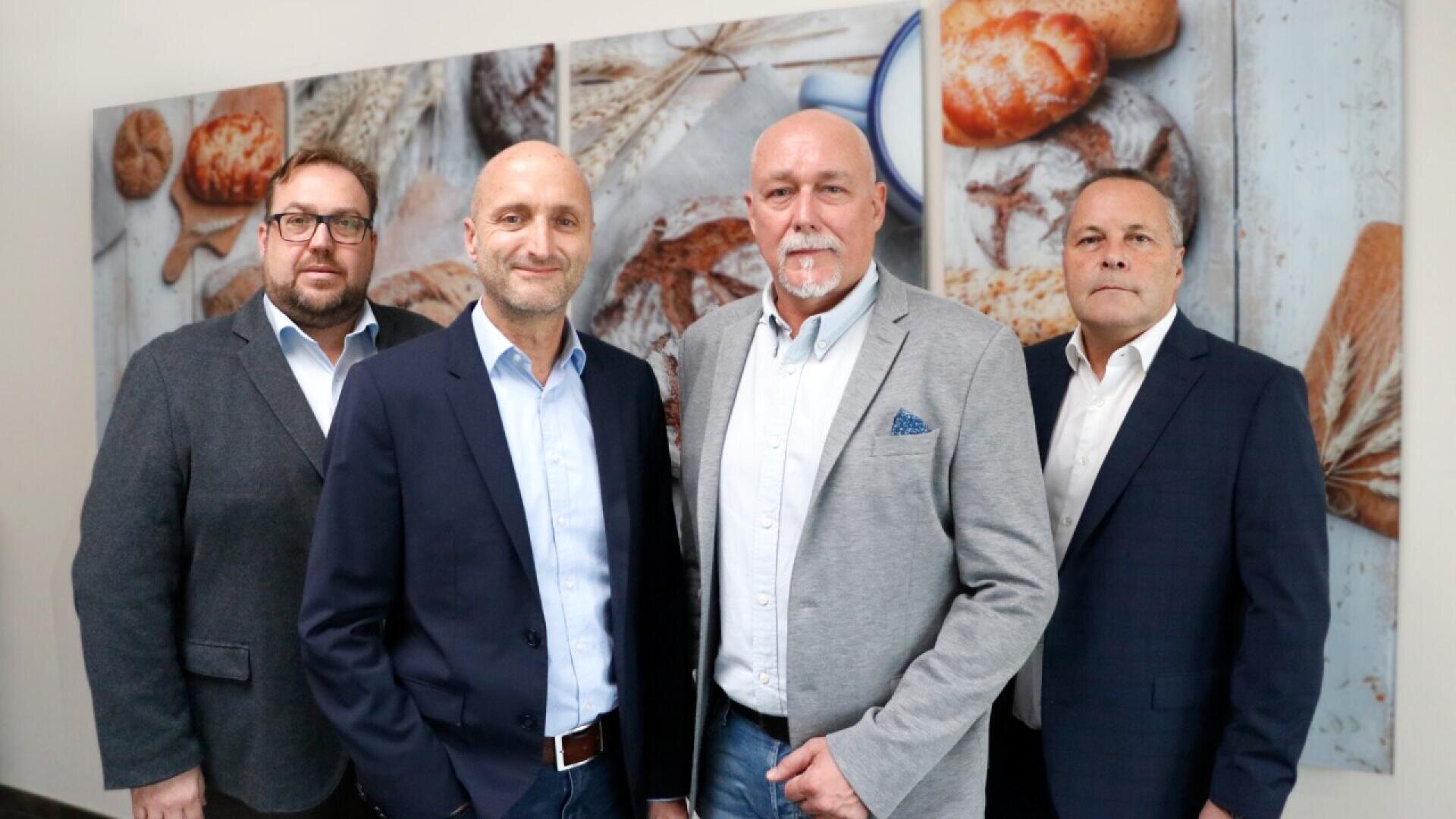 Four men in business attire stand side by side in front of a wall with large images of bread and wheat, posing for a group photo.