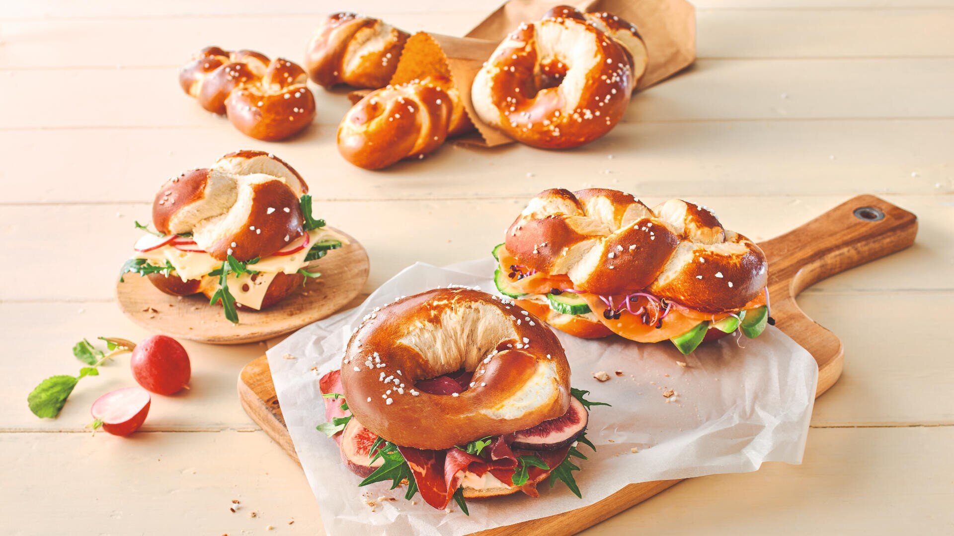 Three pretzel sandwiches filled with cheese, meats, and greens are placed on a wooden board and plate. In the background, several plain soft pretzels and rolls are arranged on a light wooden surface.