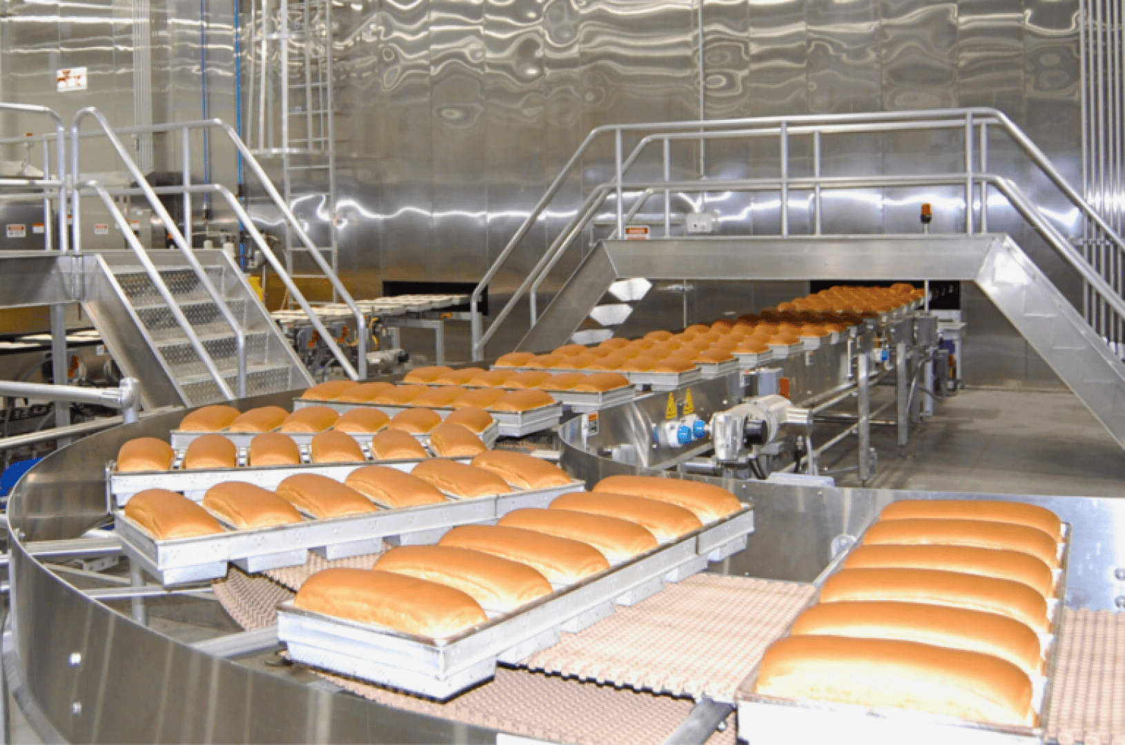 Rows of freshly baked loaves of bread move along a conveyor belt in a large, modern industrial bakery with stainless steel equipment and platforms in the background.
