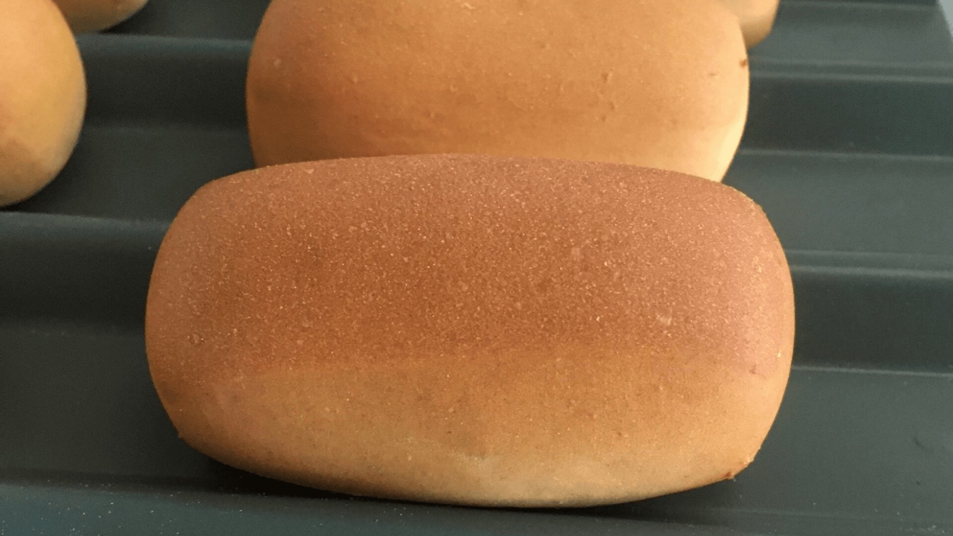 A close-up of a golden-brown rectangular bread roll on a dark baking tray, with other similar rolls visible in the background.
