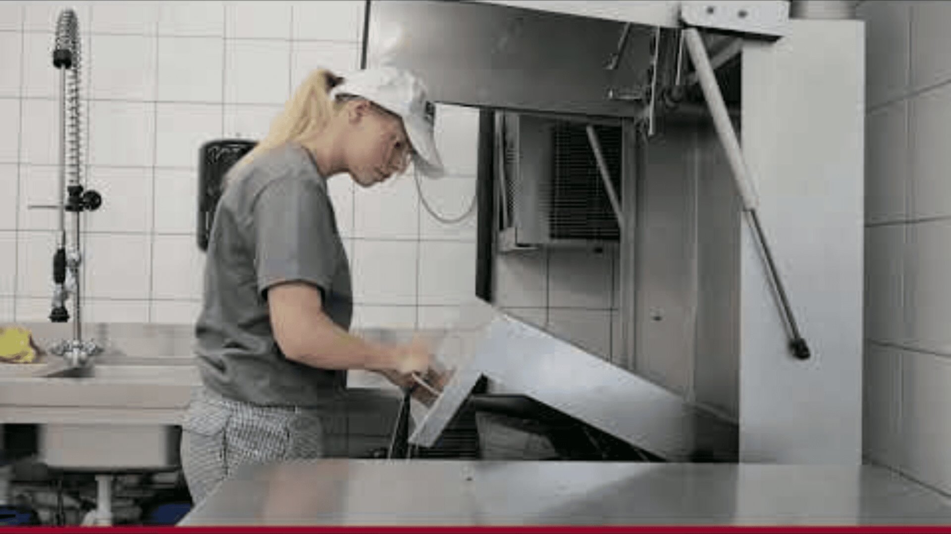 A woman wearing a cap and uniform loads a large tray into an industrial dishwasher in a commercial kitchen with white tiled walls.
