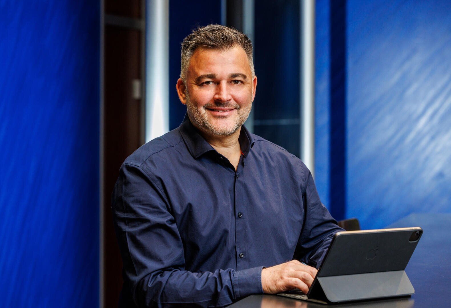 Ein Mann mit kurzen grauen Haaren und einem Bart lächelt, während er an einem Tisch sitzt, ein dunkelblaues Hemd trägt und ein Tablet benutzt. Im Hintergrund sind blaue Wände und ein modernes Interieur zu sehen.