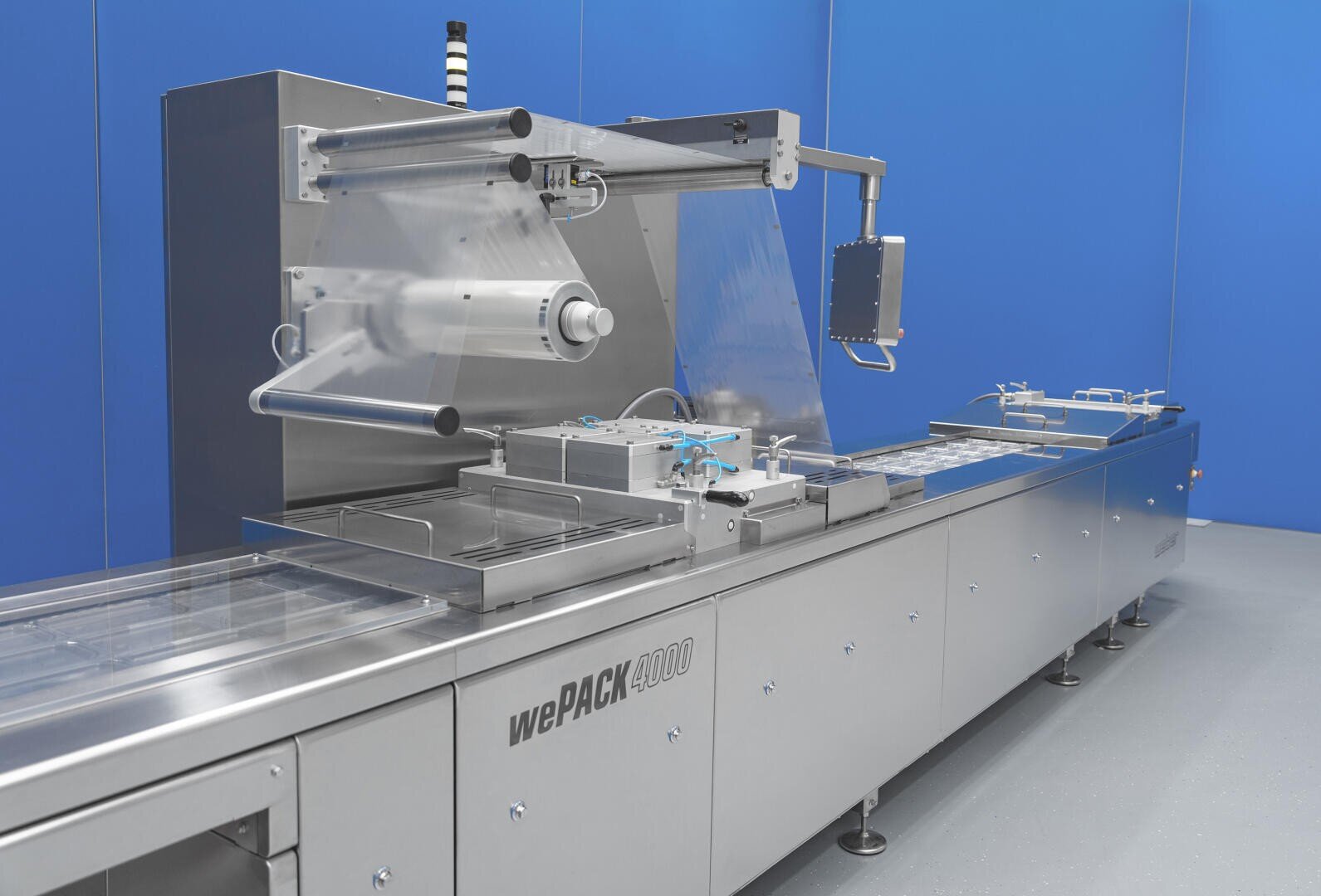 Eine Verpackungsmaschine aus Edelstahl mit der Bezeichnung wePACK 3000 steht in einem sauberen Raum mit blauen Wänden und ist mit Förderbändern, Rollen und einem Folienspender für automatisierte Verpackungsprozesse ausgestattet.