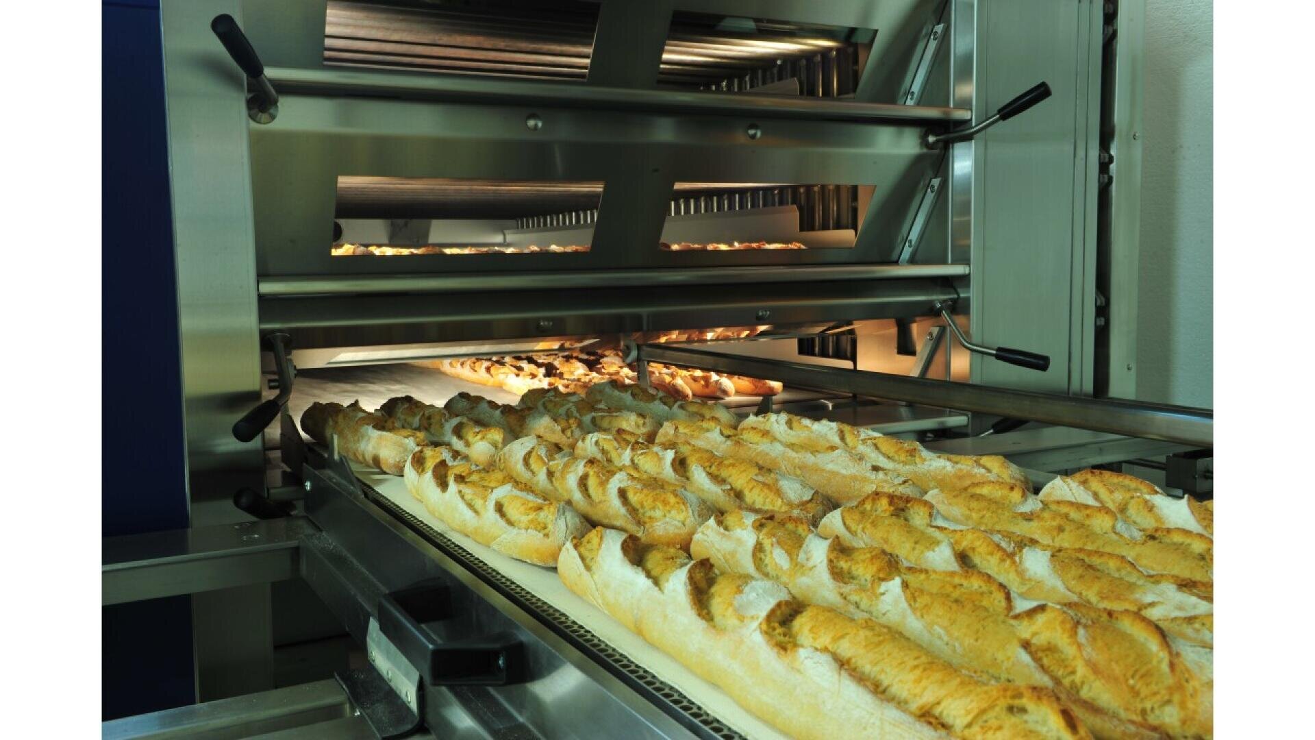 Rows of freshly baked baguettes are coming out of a large industrial oven on a conveyor belt in a commercial bakery setting.
