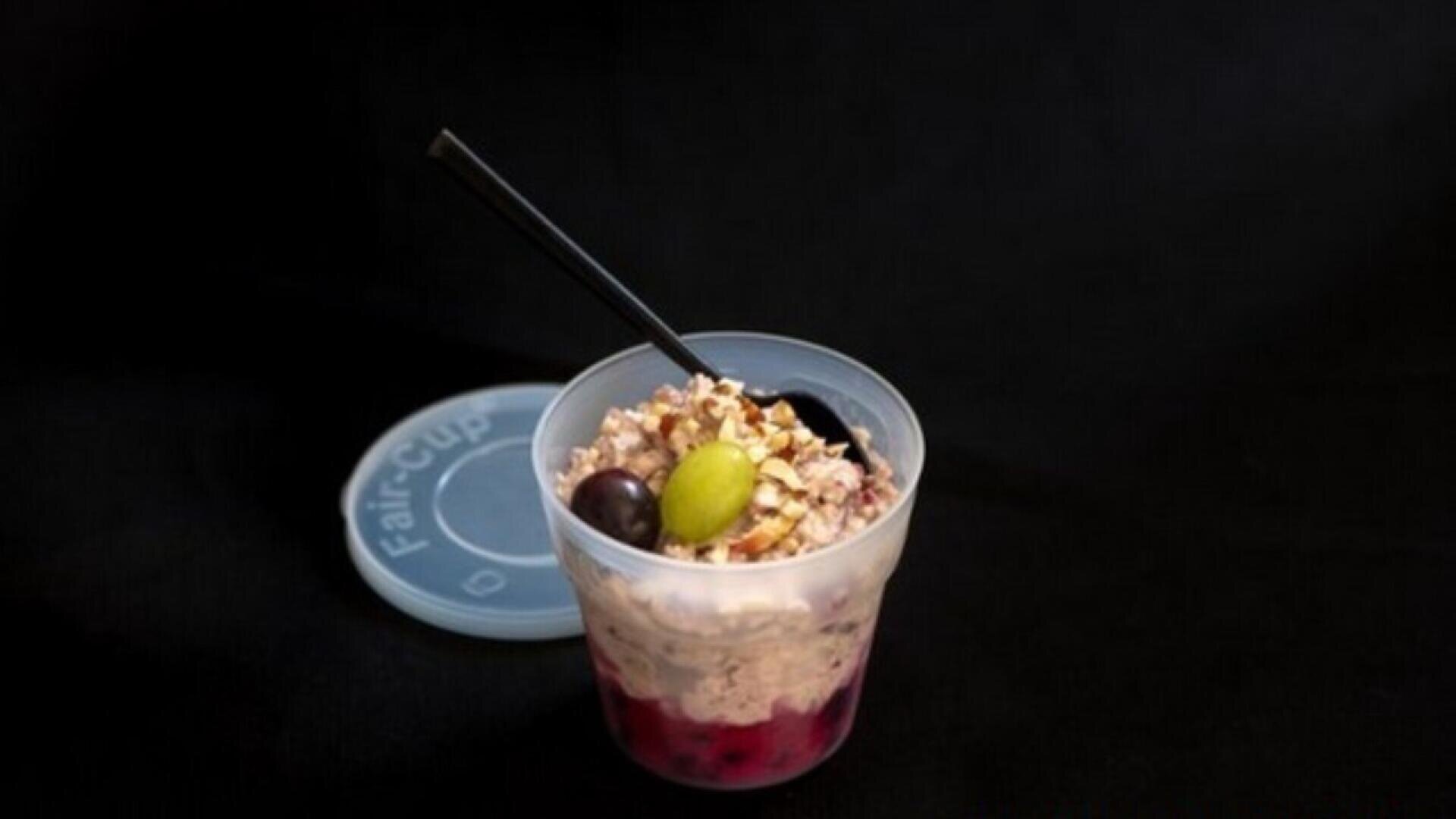 A plastic cup filled with layered yogurt, granola, nuts, grapes, and a green berry, with a black spoon inside. The lid rests beside the cup, all set against a black background.