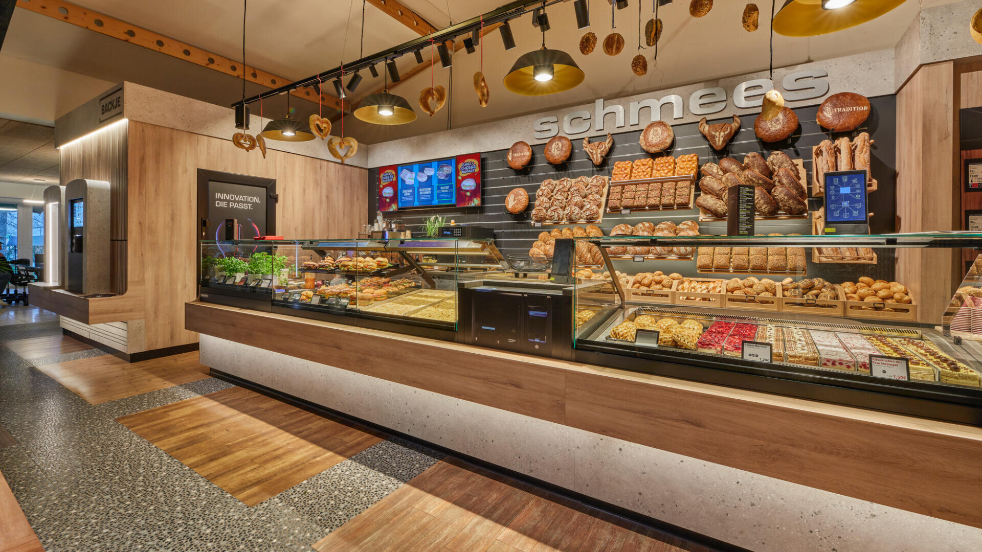 Eine moderne Bäckertheke zeigt hinter Glas verschiedene Brotsorten, Brötchen und Gebäck. Hängende Brezeln und warme Beleuchtung schaffen eine einladende Atmosphäre. Über der Theke sind digitale Menütafeln zu sehen. Der Name der Bäckerei "schmees" steht an der Wand.