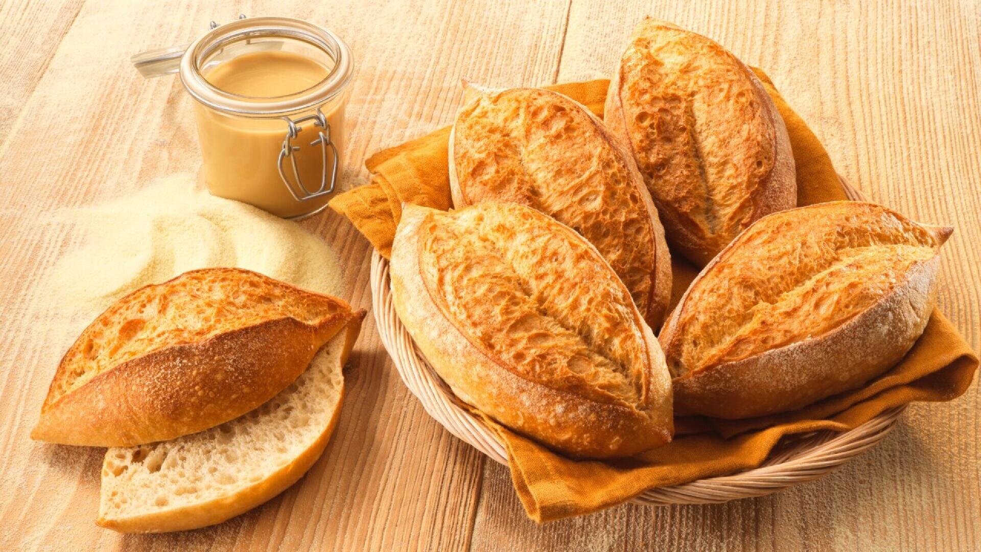 A basket with five crusty bread rolls on an orange cloth sits on a wooden table beside a cut roll, a jar of spread, and a pile of flour or semolina.