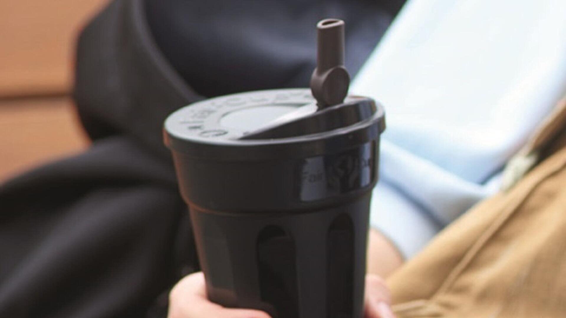 A person holds a large, black reusable cup with a raised lid and attached straw, resting on their lap. The person is wearing a tan jacket and a light blue shirt.