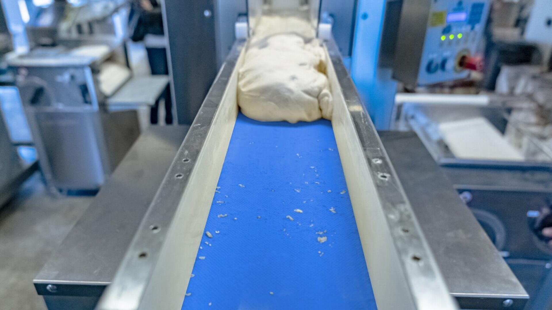 Ein Teigklumpen bewegt sich auf einem blauen Förderband in einer Lebensmittelverarbeitungsanlage, umgeben von verschiedenen industriellen Maschinen und Geräten.