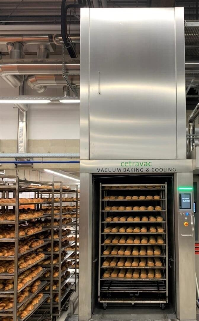 A commercial bakery with racks of baked goods and a large CetraVac vacuum baking and cooling machine filled with trays of bread rolls. Industrial pipes and lighting are visible on the ceiling above.