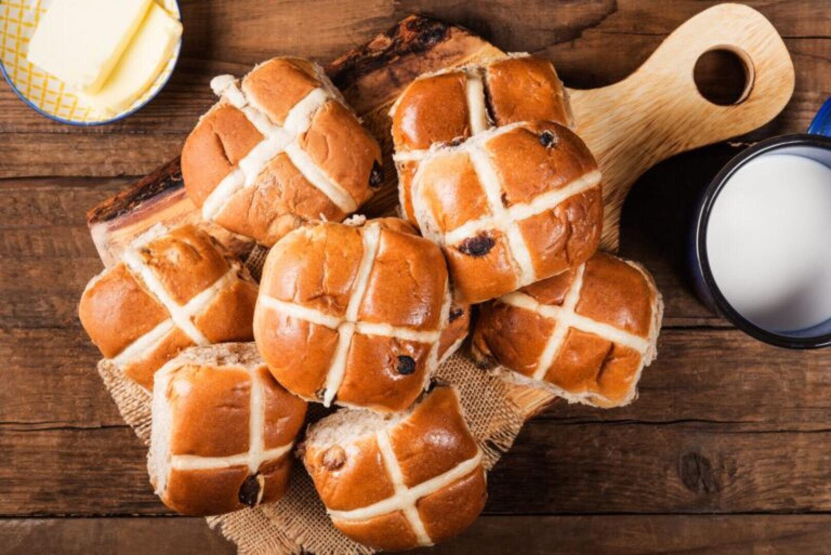 A wooden board holds a pile of hot cross buns with white icing crosses, next to a cup of milk and a dish of butter, all on a rustic wooden table.