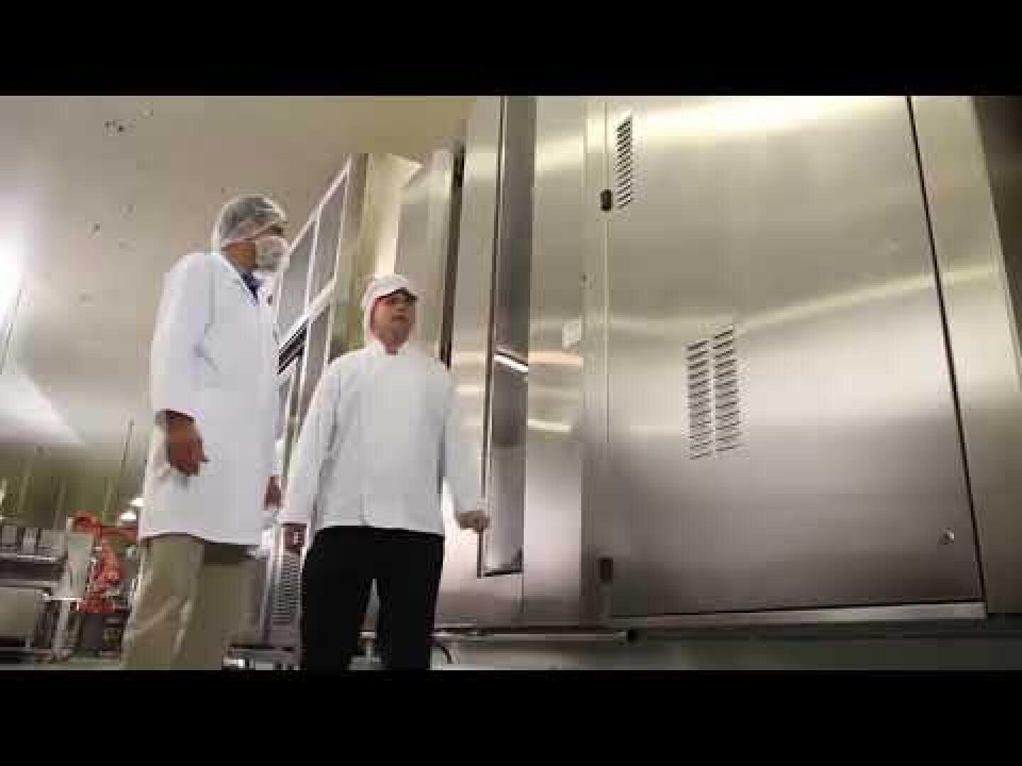 Zwei Personen in weißen Laborkitteln und Haarnetzen stehen vor großen Industriegeräten aus Edelstahl in einer sauberen, hell erleuchteten Anlage, möglicherweise einem Lebensmittel- oder Pharmabetrieb.